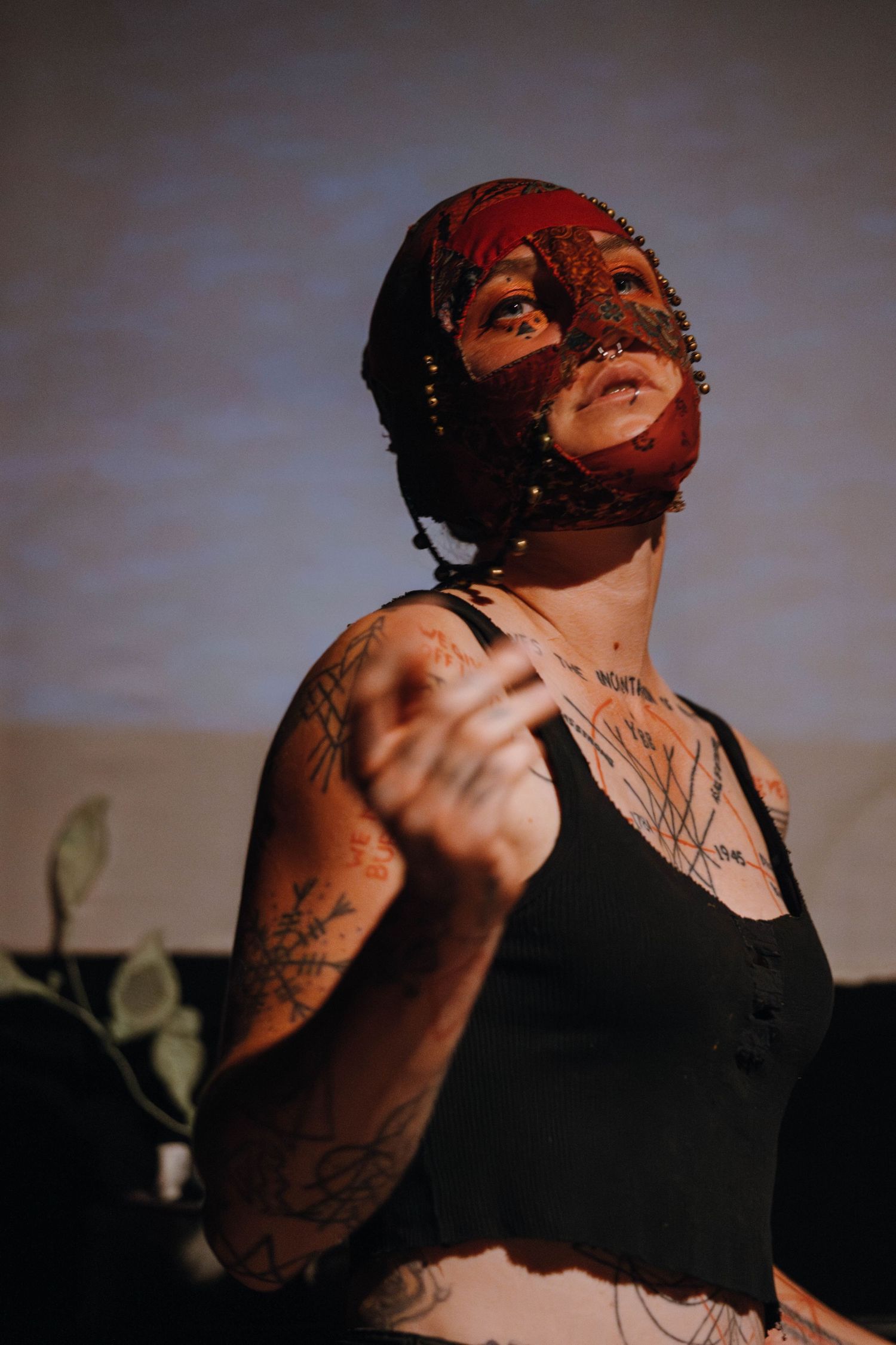 Tattooed performer wearing ornate red mask gazes upward dramatically under warm stage lighting.