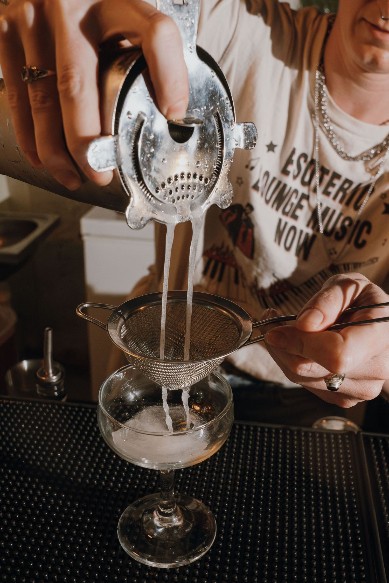 Bartender double straining a cocktail into a coupe glass at a bar.