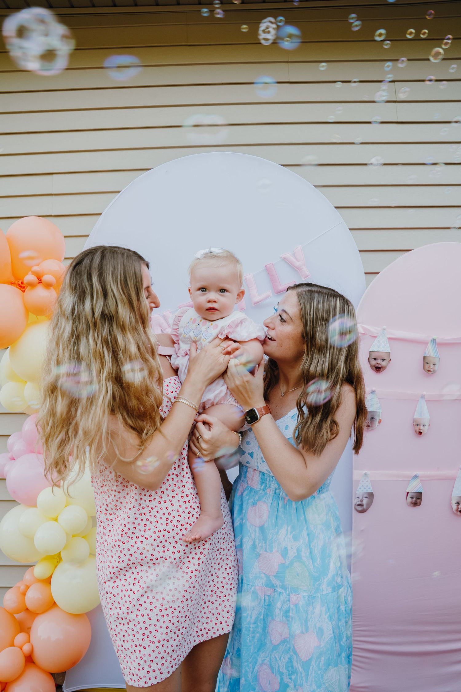 Two women holding a baby girl surrounded by colorful balloons and bubbles at a birthday party.
