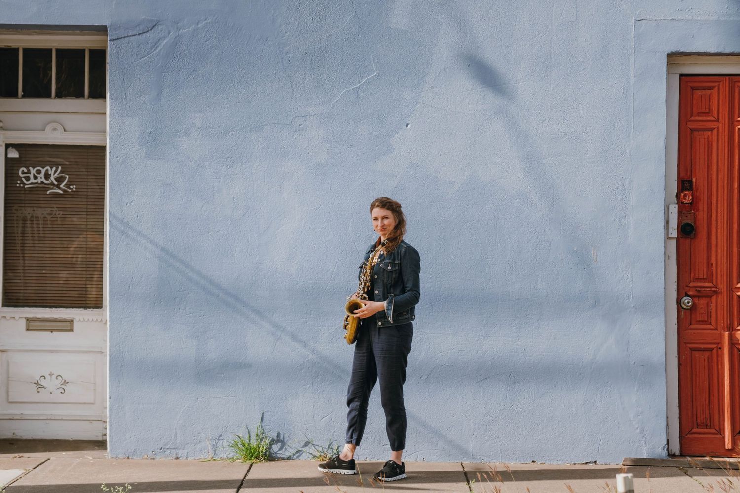 Female saxophonist standing against light blue building exterior on urban sidewalk holding saxophone.