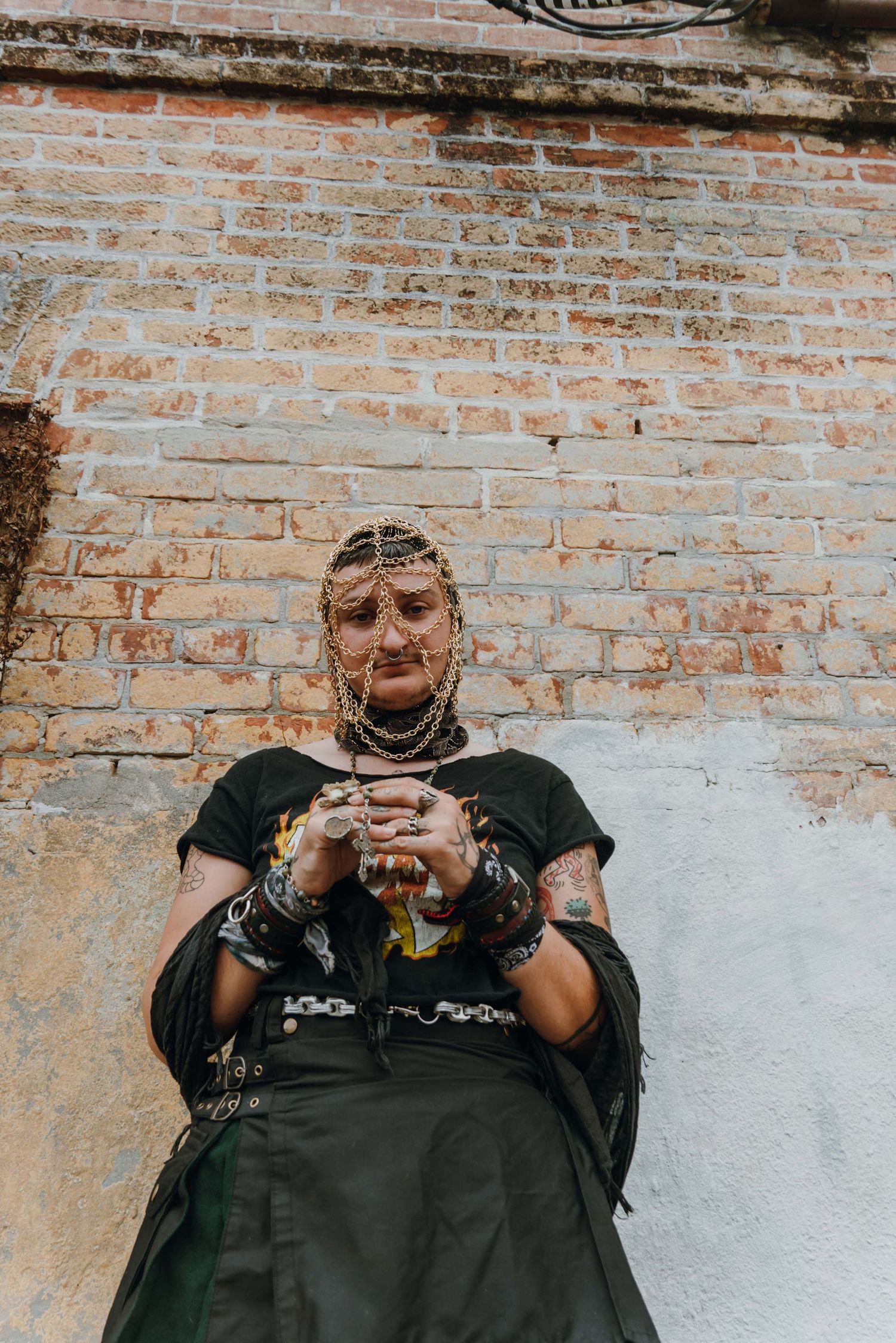 Tattooed person adorned with gold chain headpiece and jewelry leaning against a rustic brick wall.