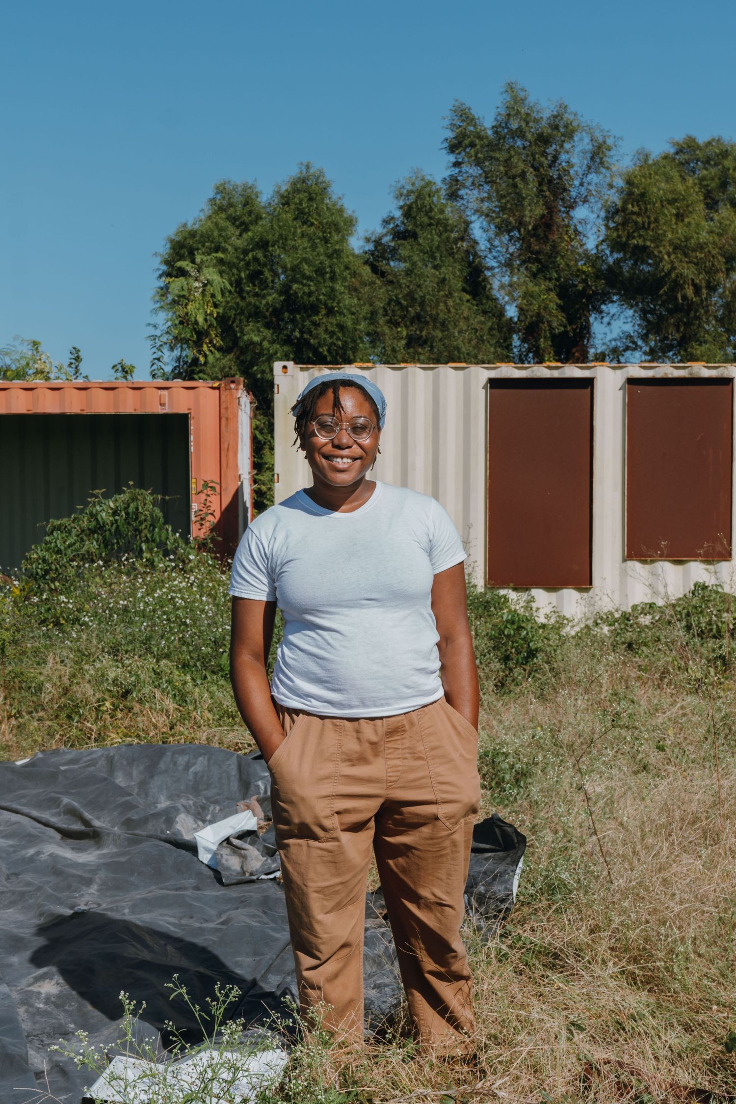 Smiling woman in glasses and brown pants standing in front of shipping containers in a sunny field.