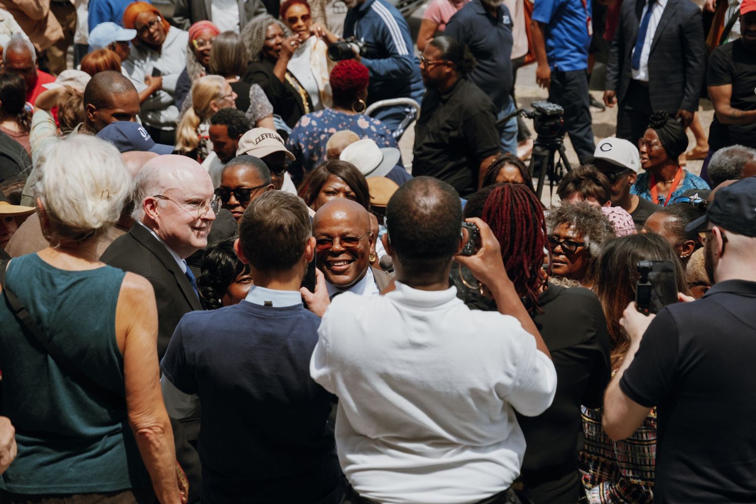 Diverse crowd of supporters surrounding a bald man in white shirt at an outdoor political campaign event.