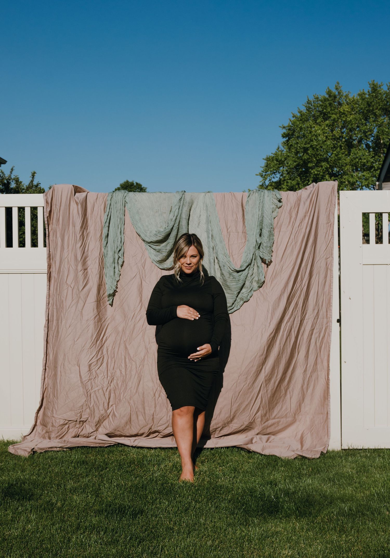 Pregnant woman in black dress walking barefoot on grass with fabric backdrop outdoors.
