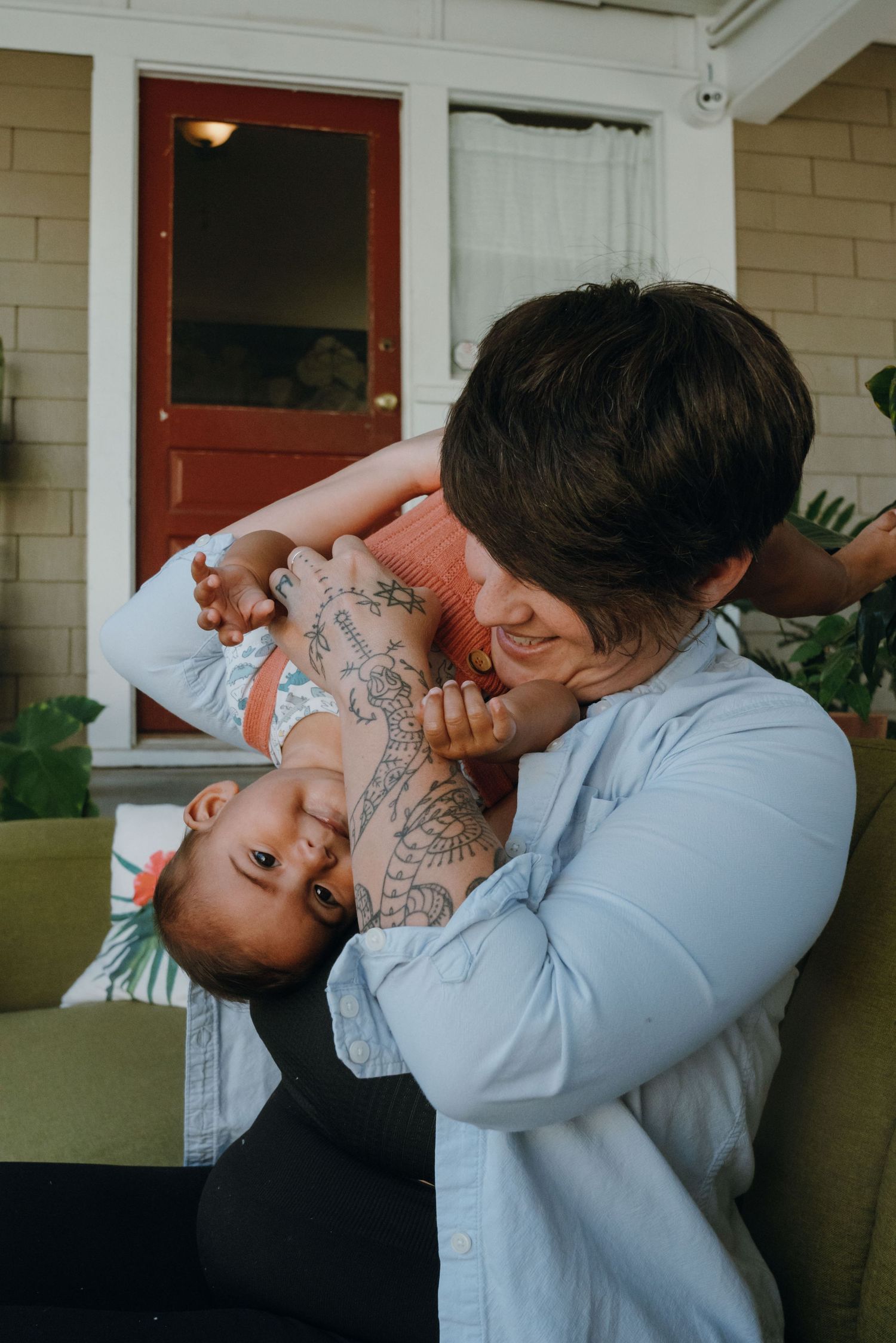 Tattooed parent smiling while playfully holding a laughing baby upside down on a home porch.