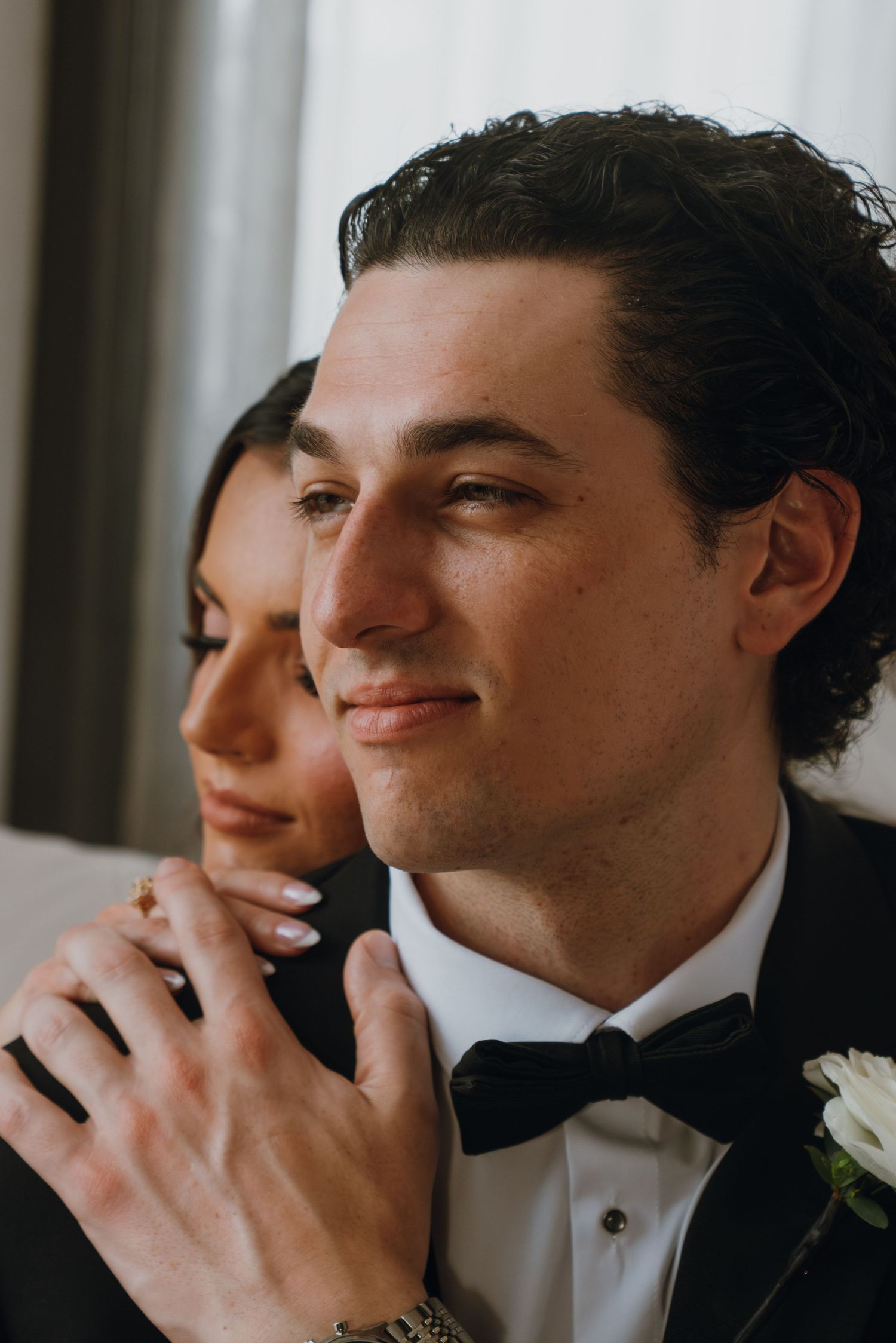 Groom in black tuxedo smiling with bride resting her hands on his shoulders in an intimate wedding portrait.