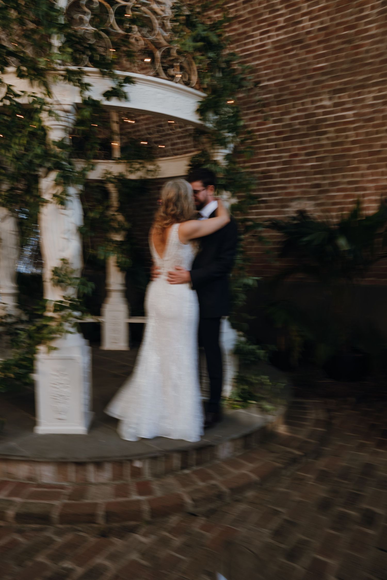 Bride and groom share a romantic embrace near ivy-covered white columns on a brick patio.