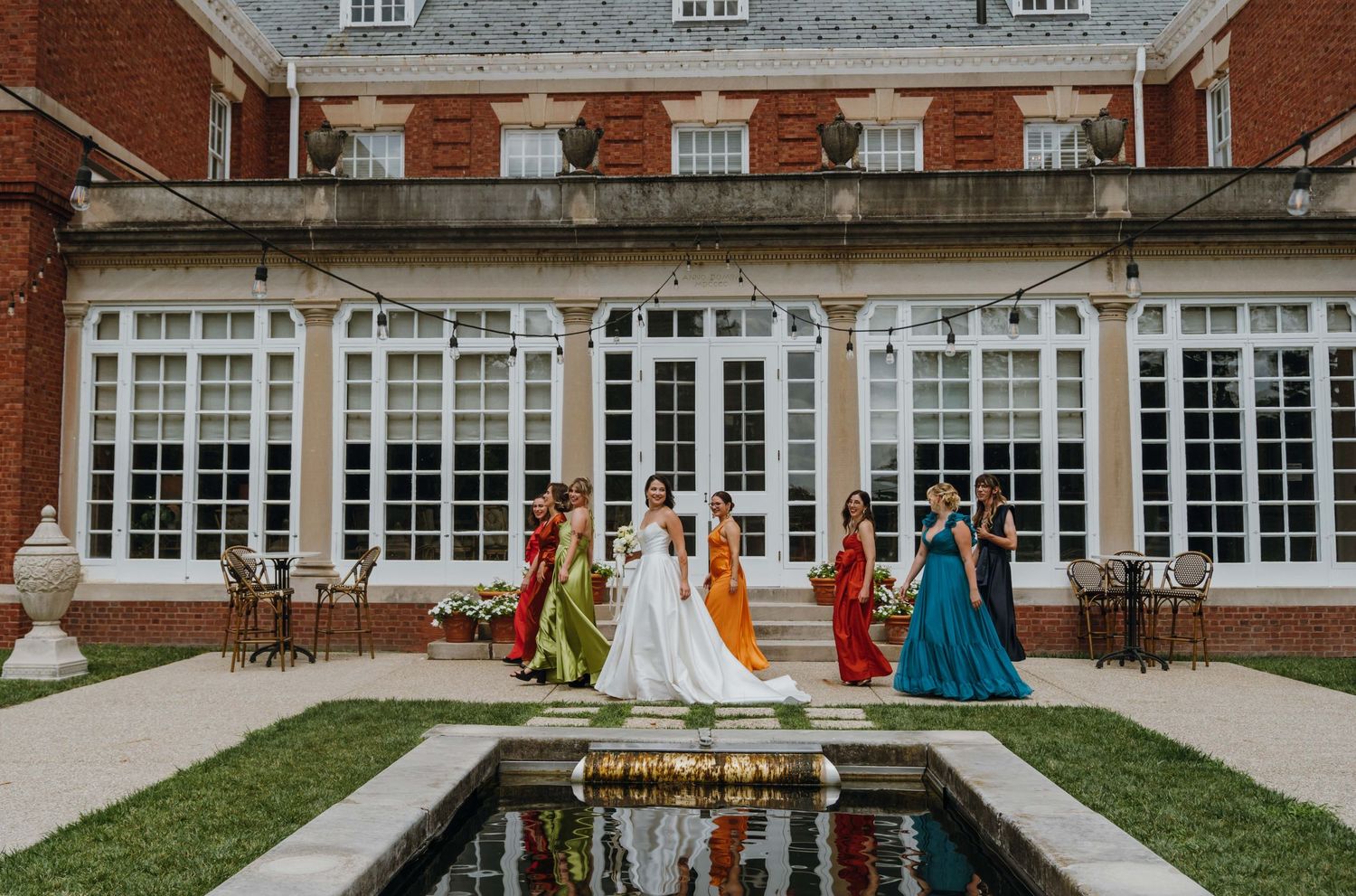 Bride and colorful bridesmaids pose by a reflecting pool outside a grand brick manor. New Orleans, Louisiana, Mississippi