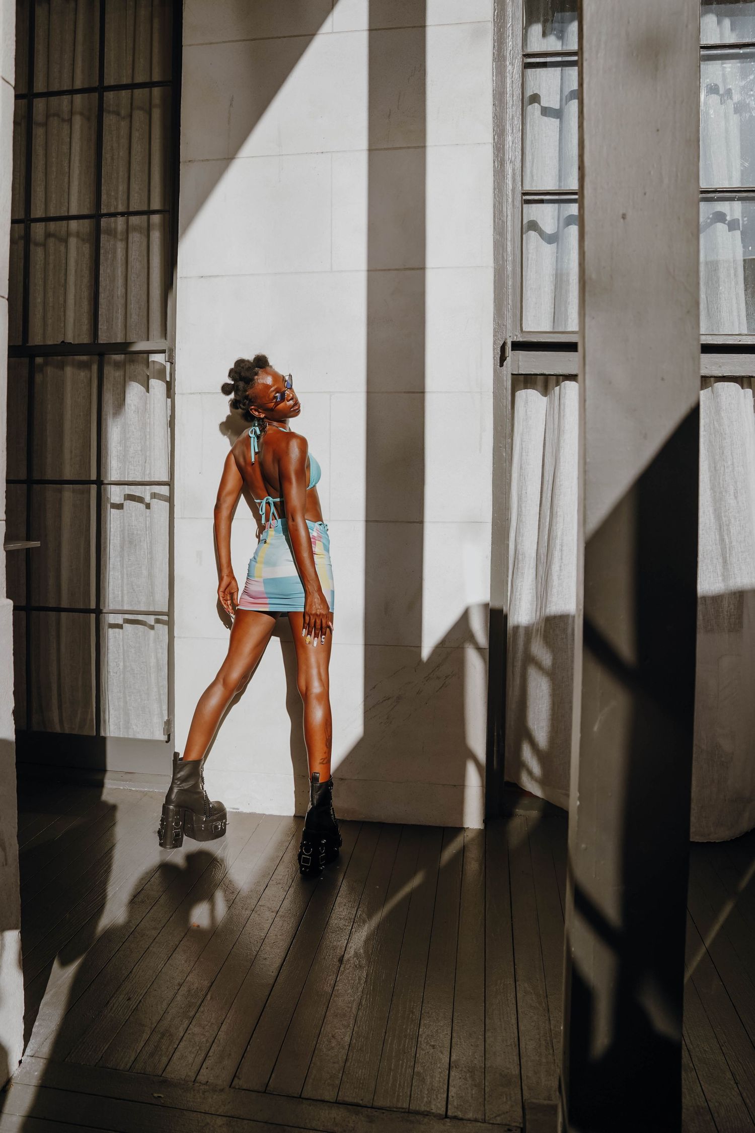 A young woman in a colorful mini dress poses in dramatic sunlight streaming through tall windows.