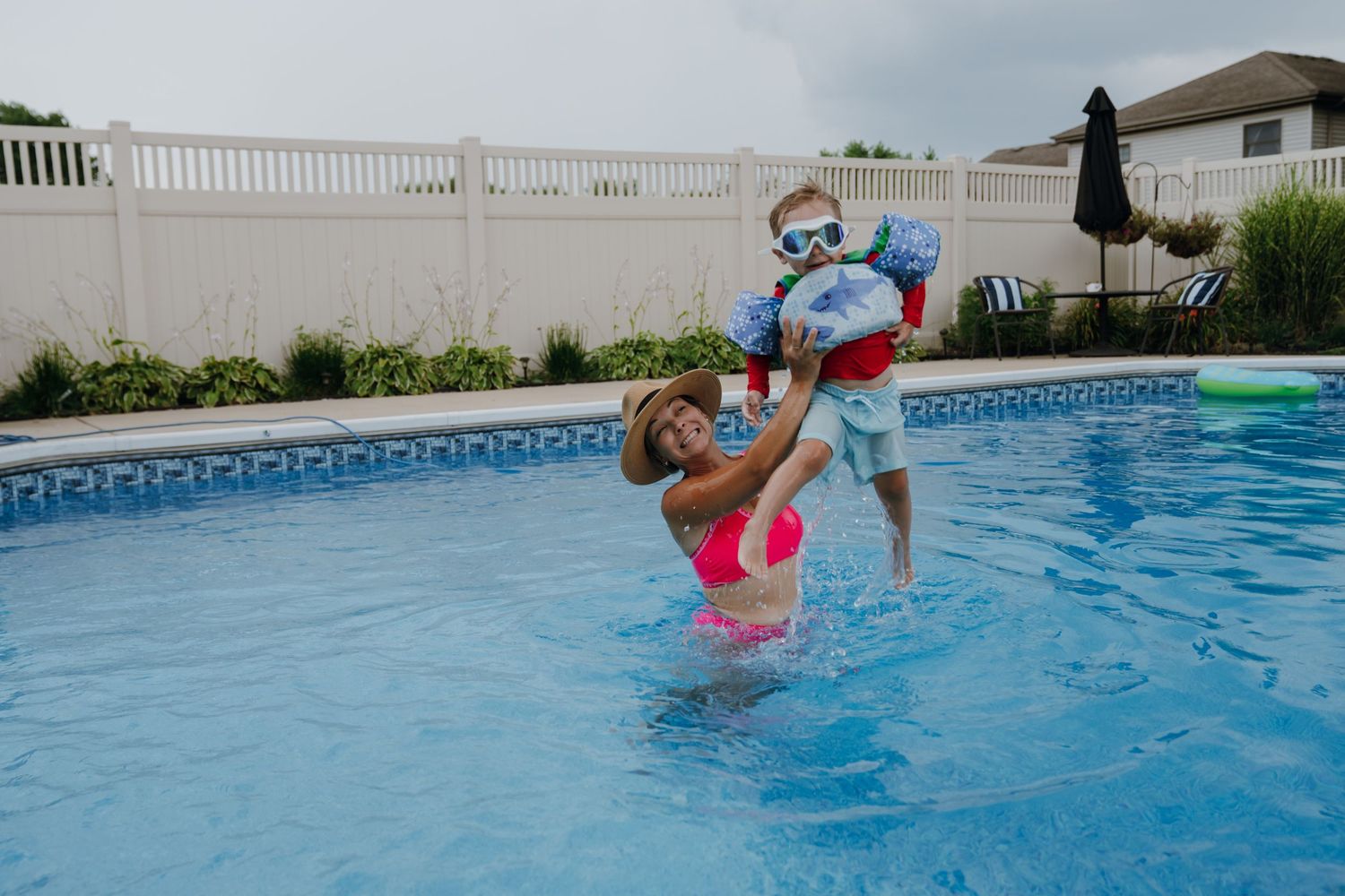 Mother lifting young child with floaties in a backyard swimming pool on a cloudy day.
