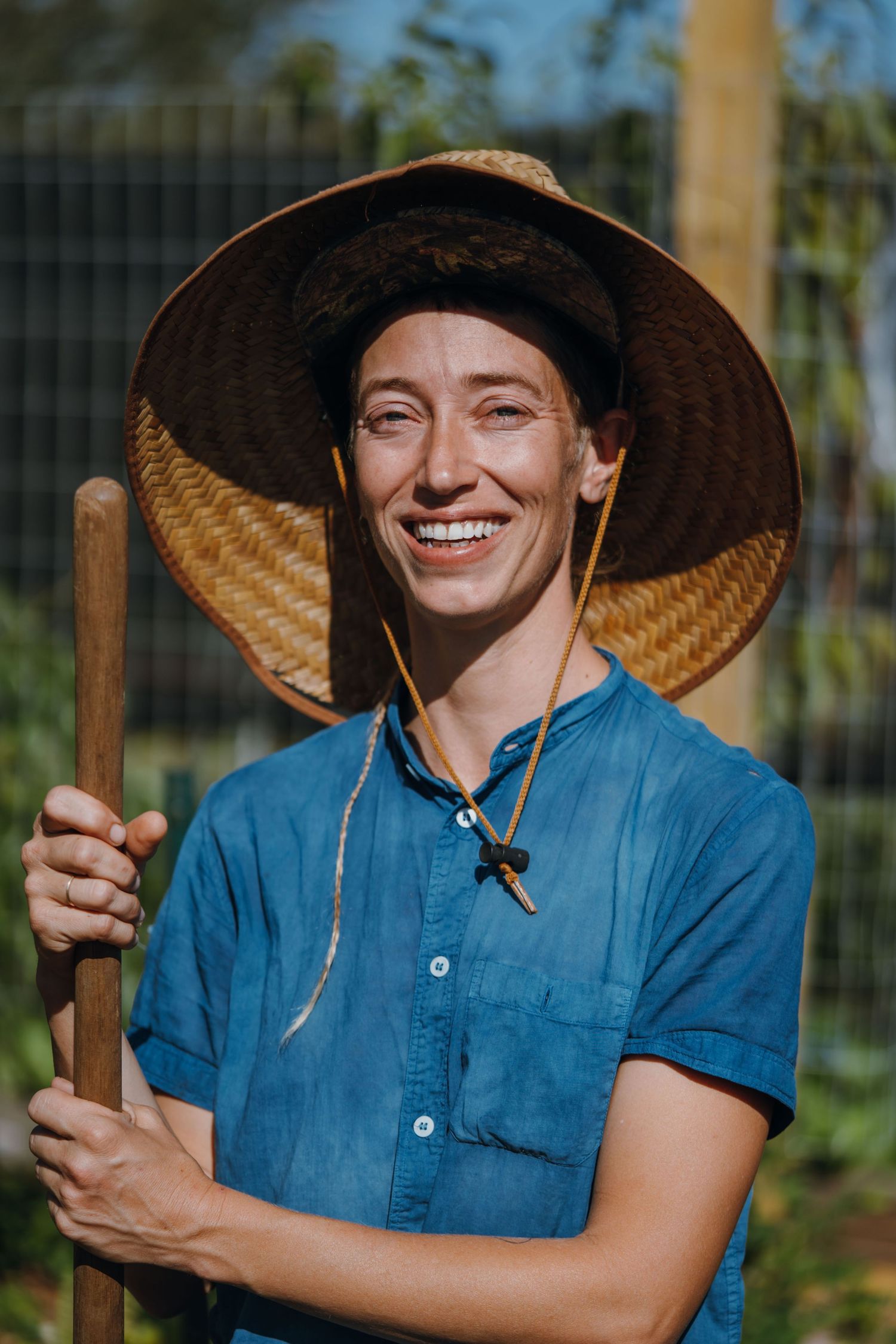 Cheerful person in a wide straw hat and blue shirt holding a wooden garden tool outdoors.