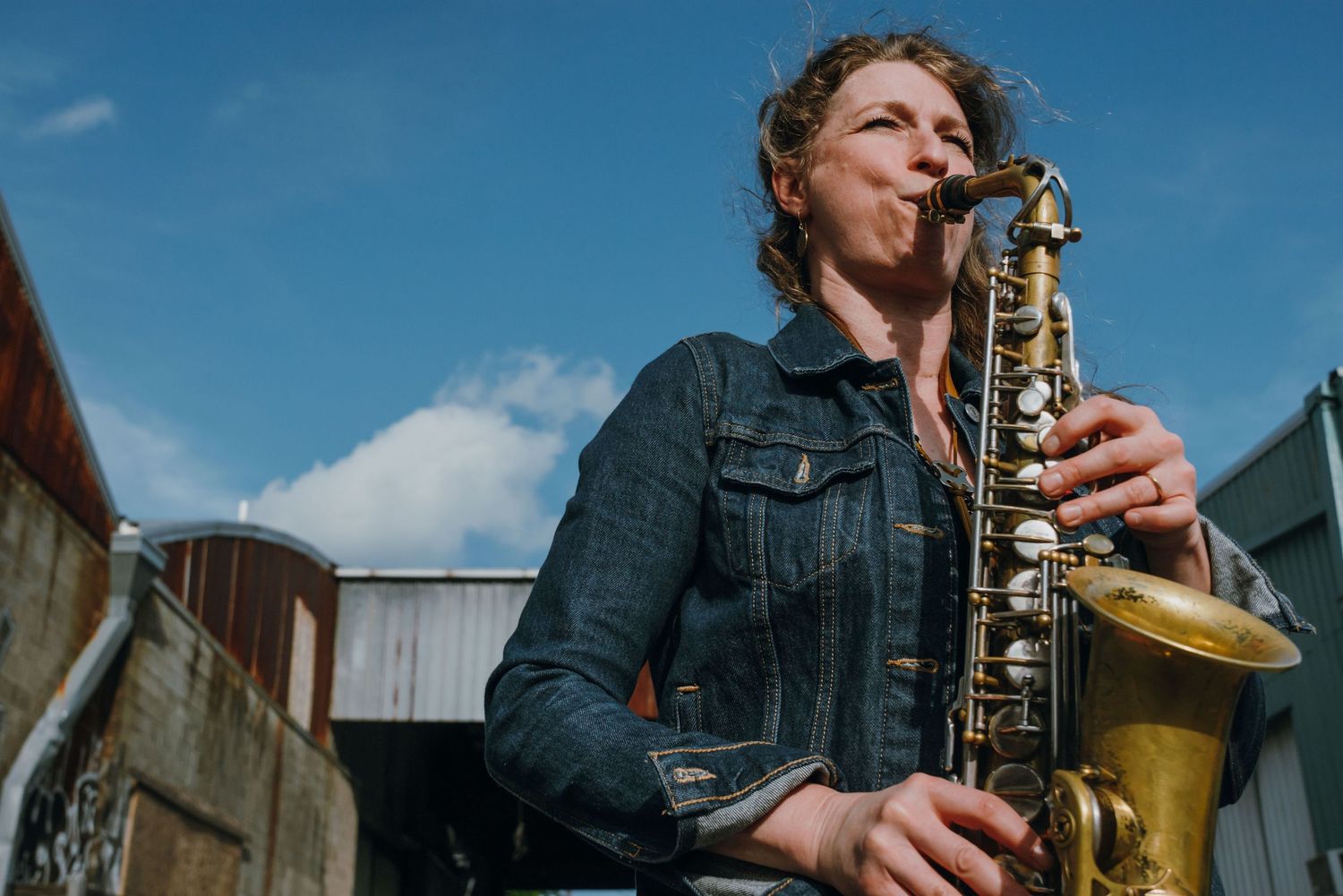 Female saxophonist playing brass saxophone outdoors against blue sky wearing denim jacket.