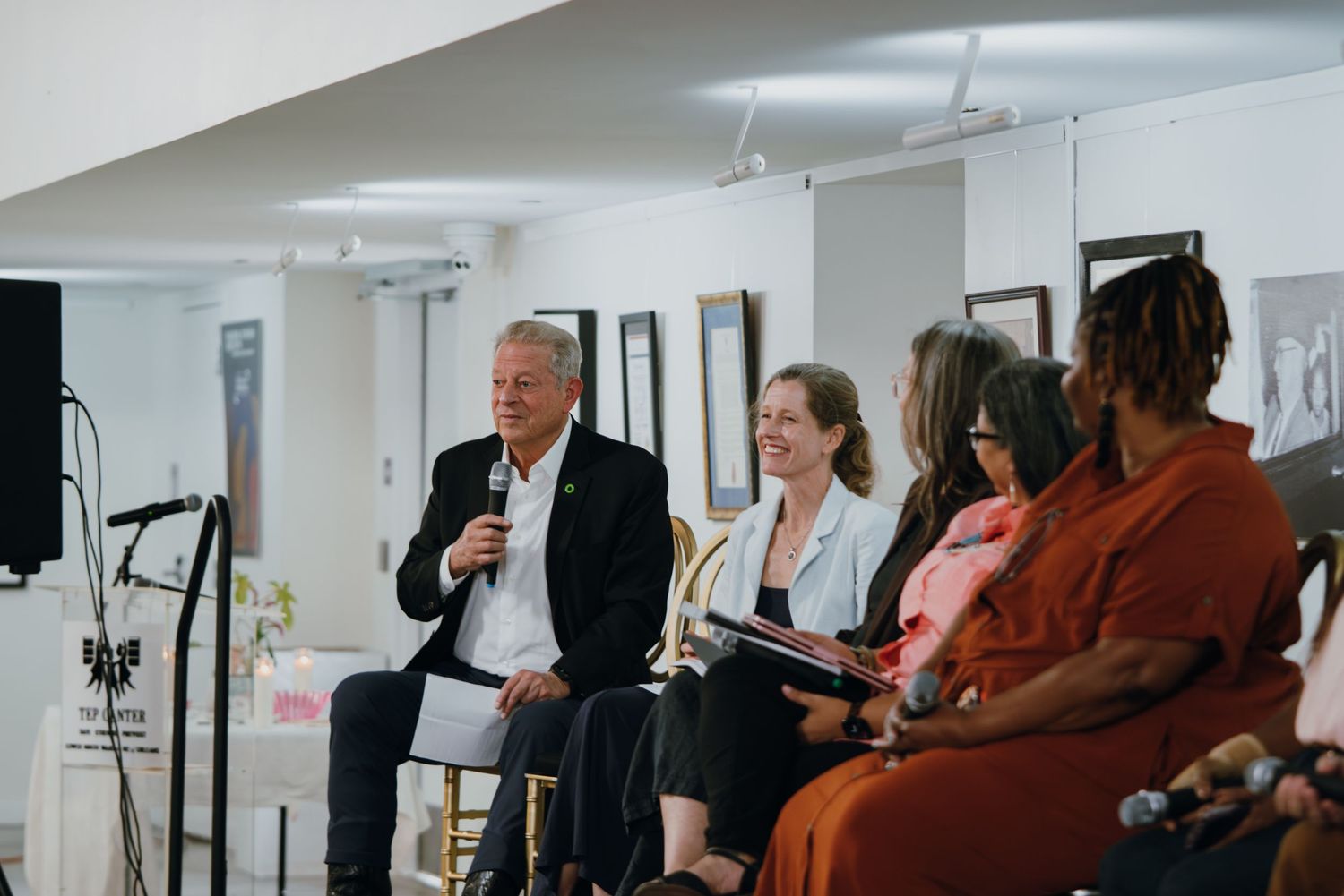 A diverse panel discussion with speakers seated in chairs, one man speaking into a microphone at a community event.