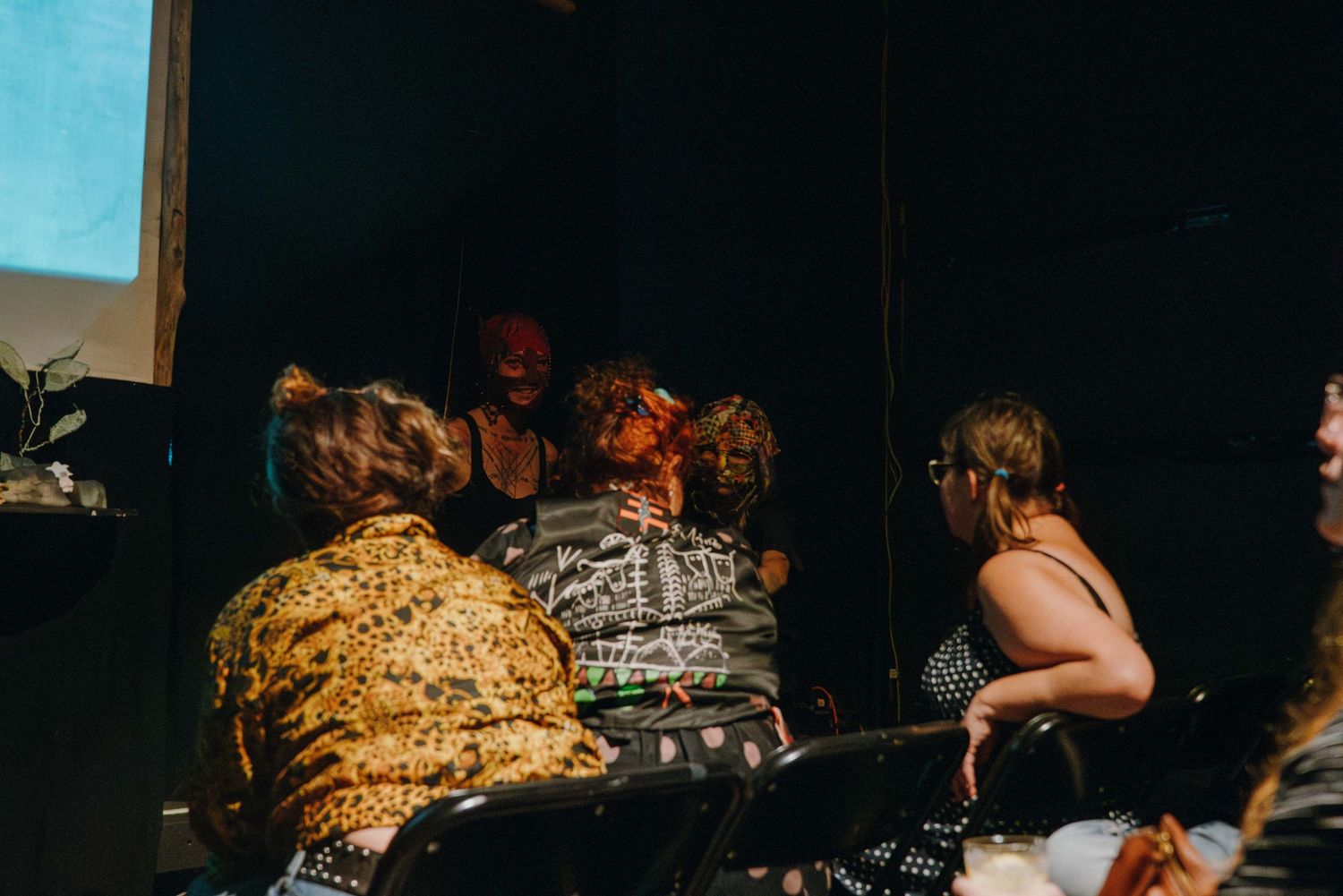 Masked performers interact with seated audience members in an intimate dark theater setting.