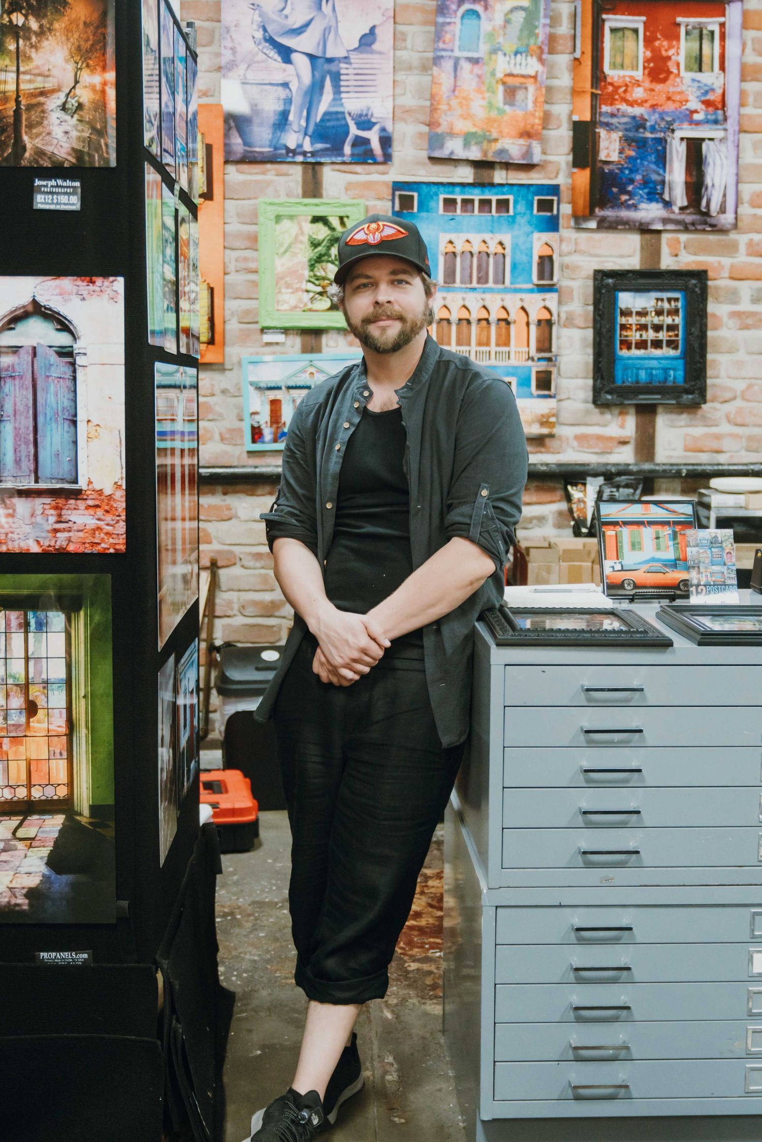 Man in black outfit standing in front of colorful urban photography prints in an art studio.
