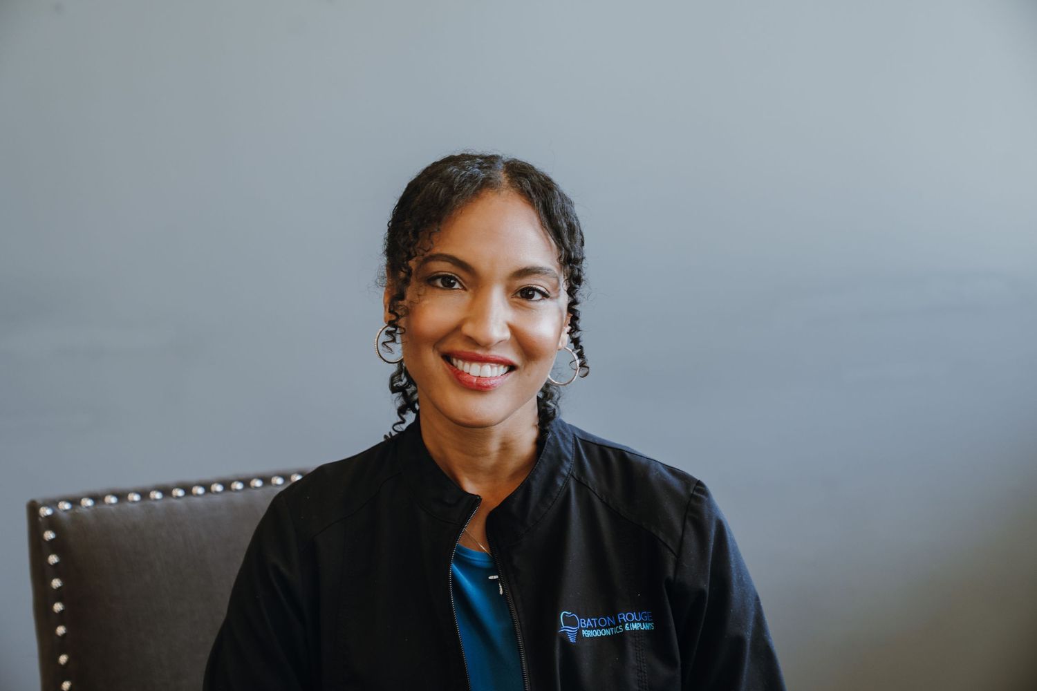 A smiling woman in a black dental jacket poses professionally against a gray background.