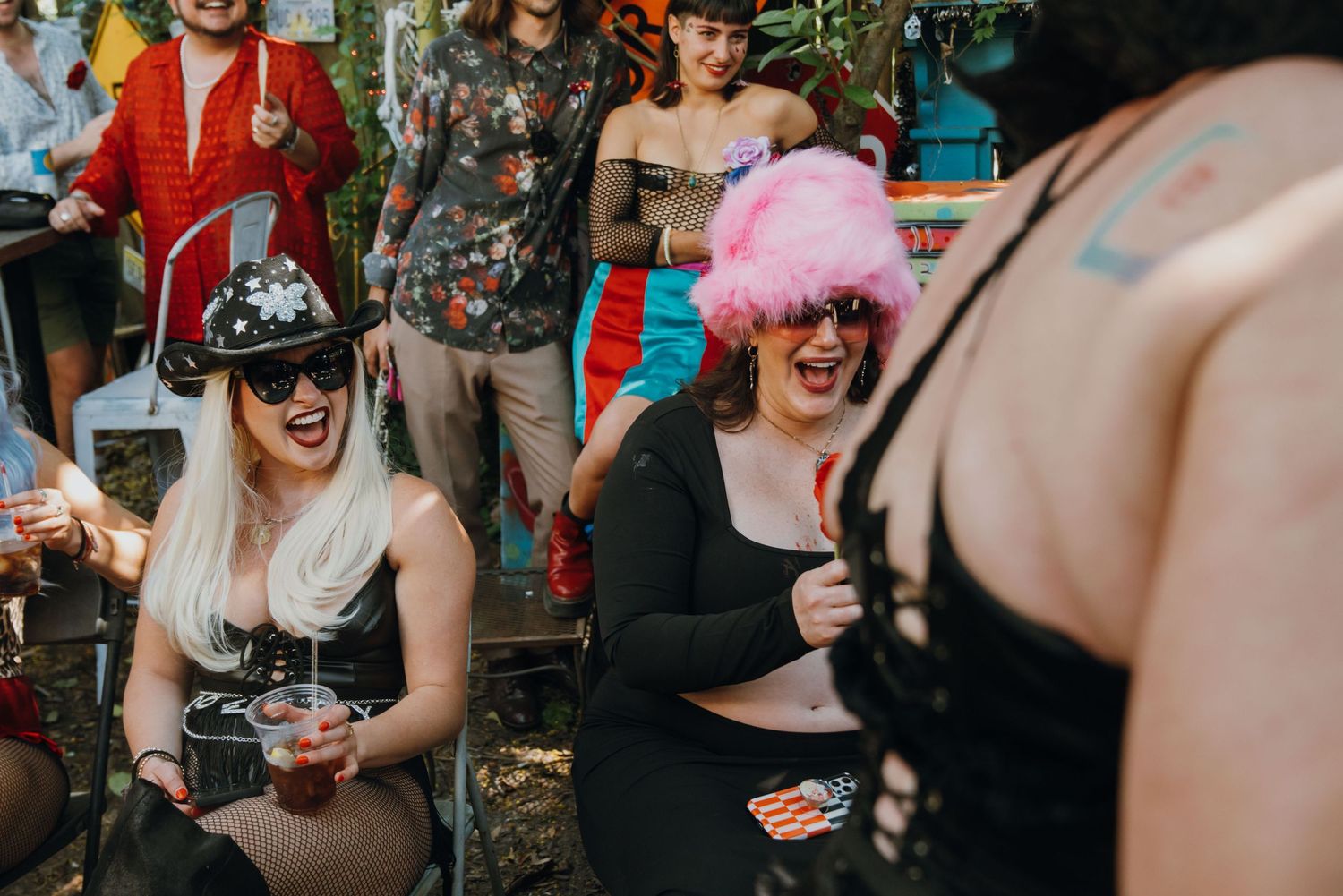 Two women in fun wigs and festive outfits laugh together at an outdoor party gathering.