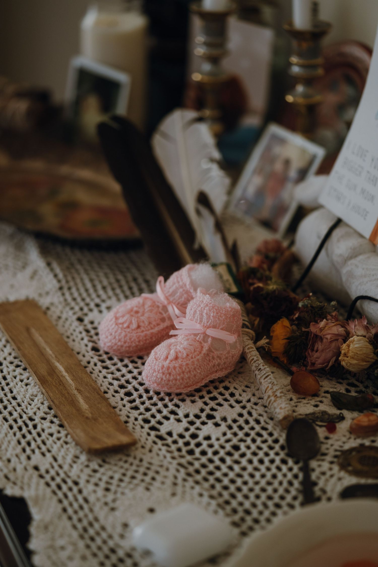 Tiny pink crocheted baby booties with bow on lace tablecloth surrounded by candles and dried flowers.