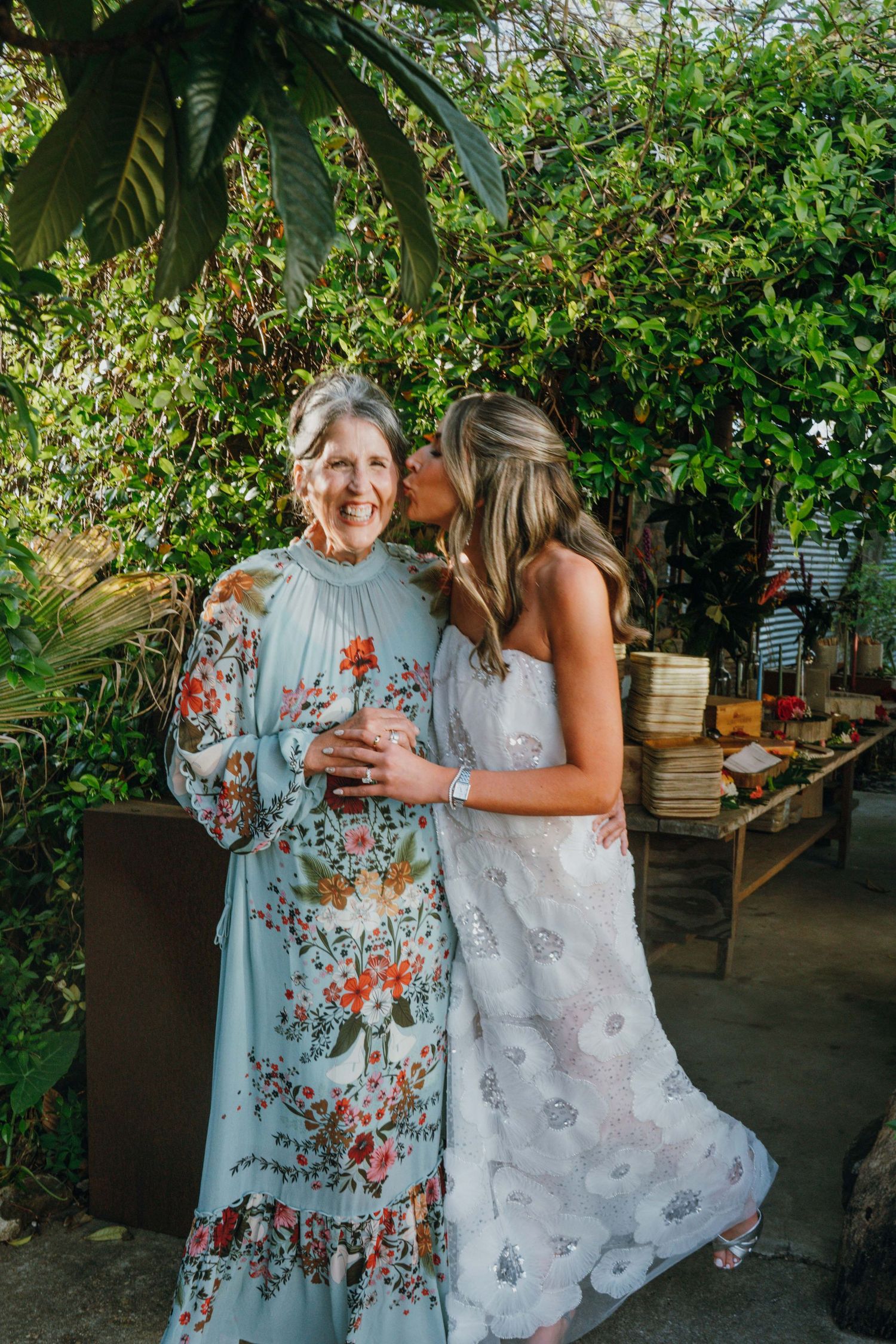 Bride in white floral gown kisses older woman in blue floral dress on the cheek at an outdoor garden wedding.