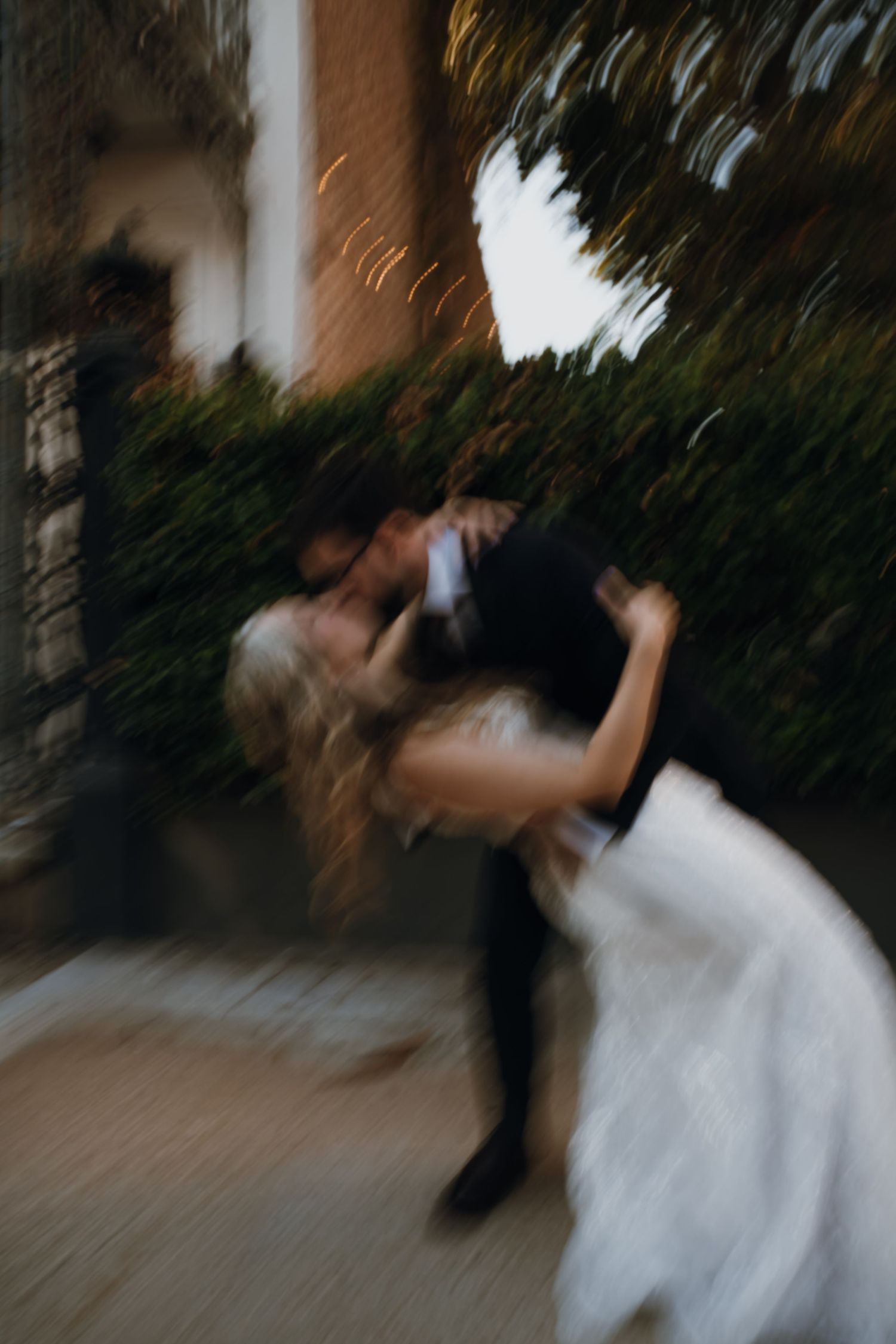 Groom dips bride in a dramatic kiss outside a brick building with green foliage.