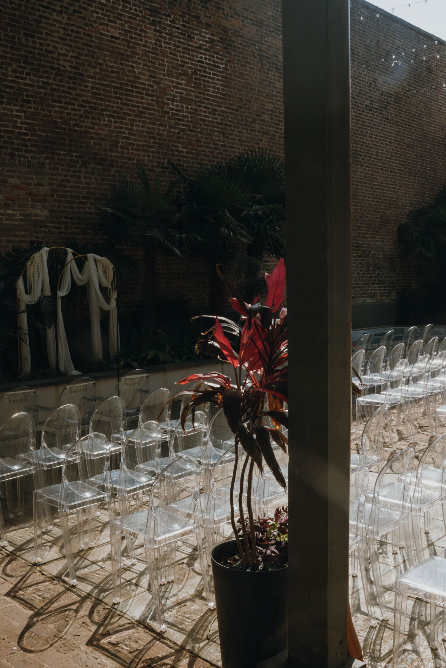 Outdoor wedding ceremony setup with ghost chairs, red tropical plants, and brick wall backdrop in sunlight.