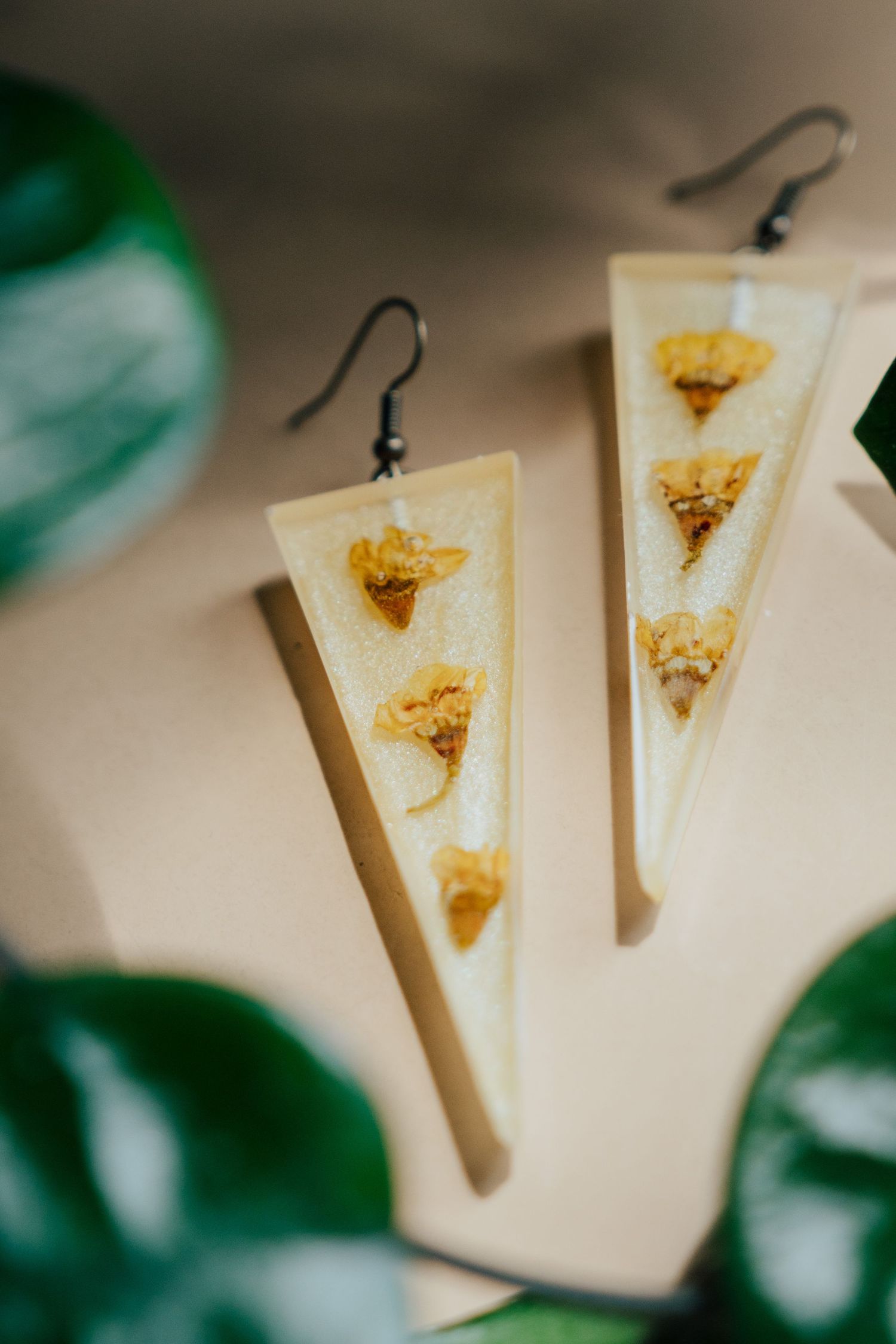 Handmade triangular resin earrings with dried yellow flowers suspended inside, displayed near green leaves.