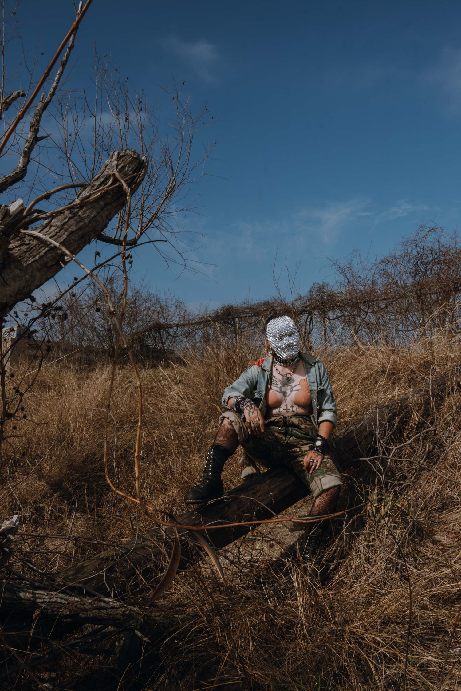Person wearing a white mask sitting on a log in a dry grassland field under blue sky.
