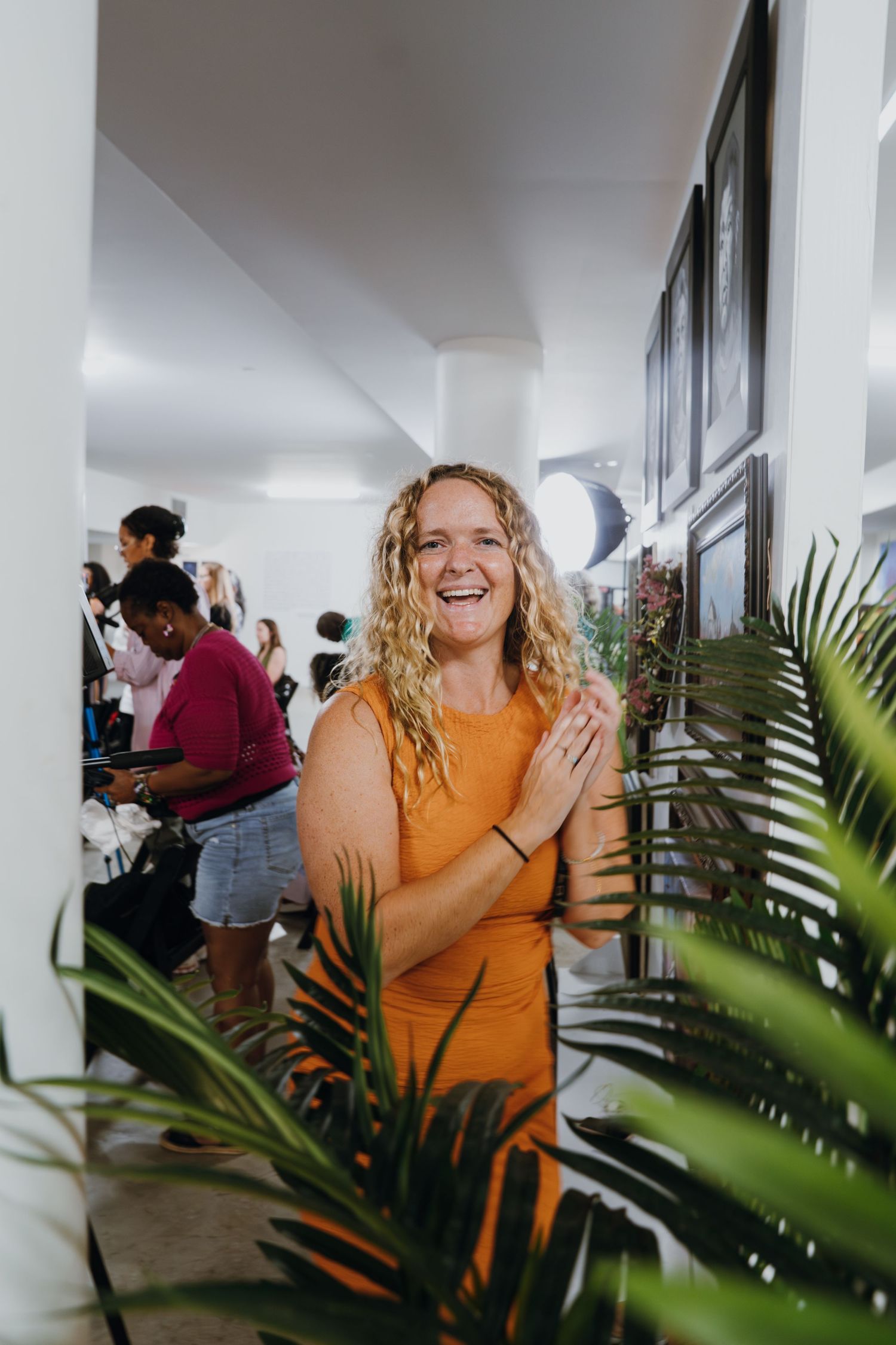 A smiling blonde woman in an orange dress stands near tropical plants at an indoor community gathering.