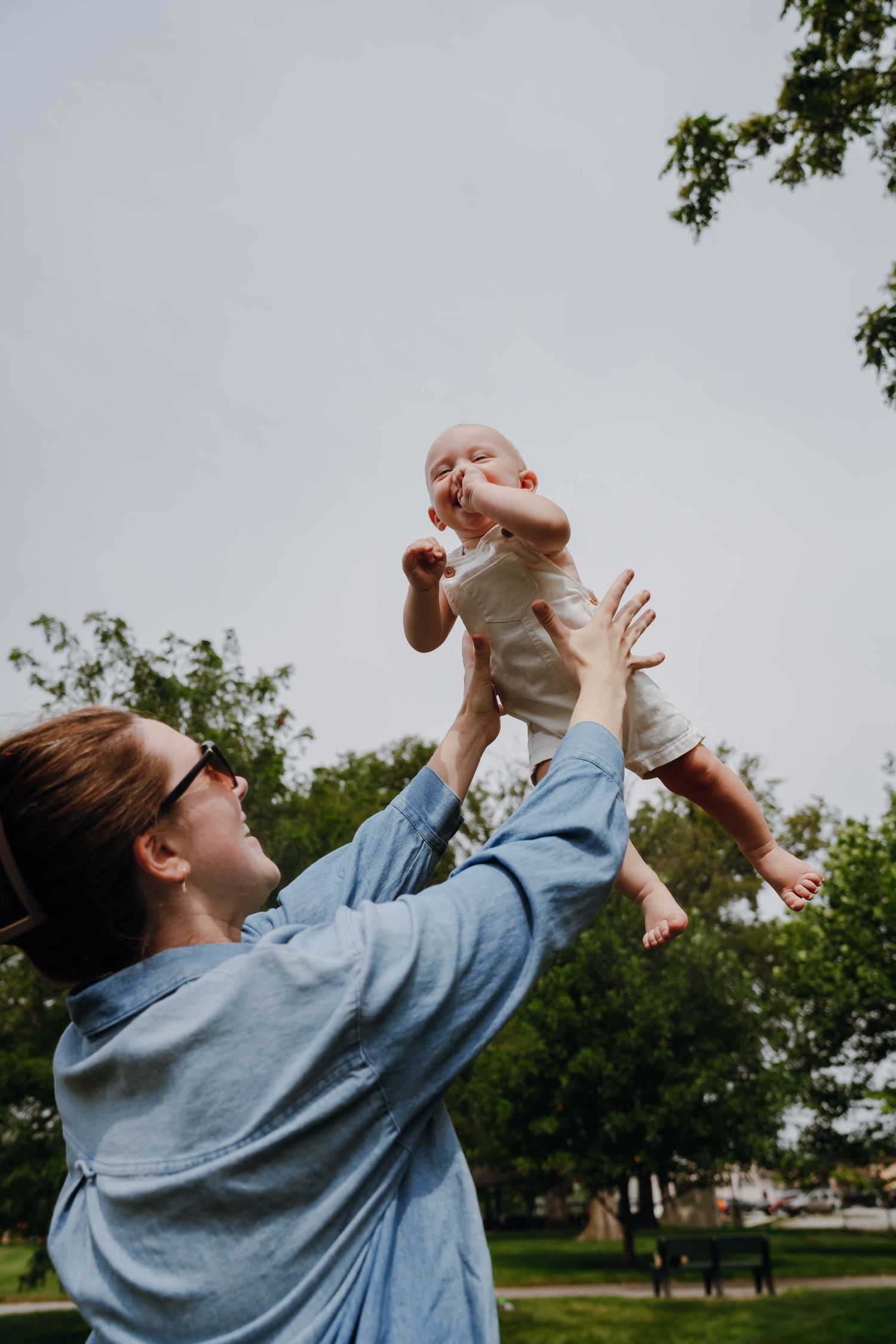 Mother lifting smiling baby in the air at an outdoor park on a cloudy day.
