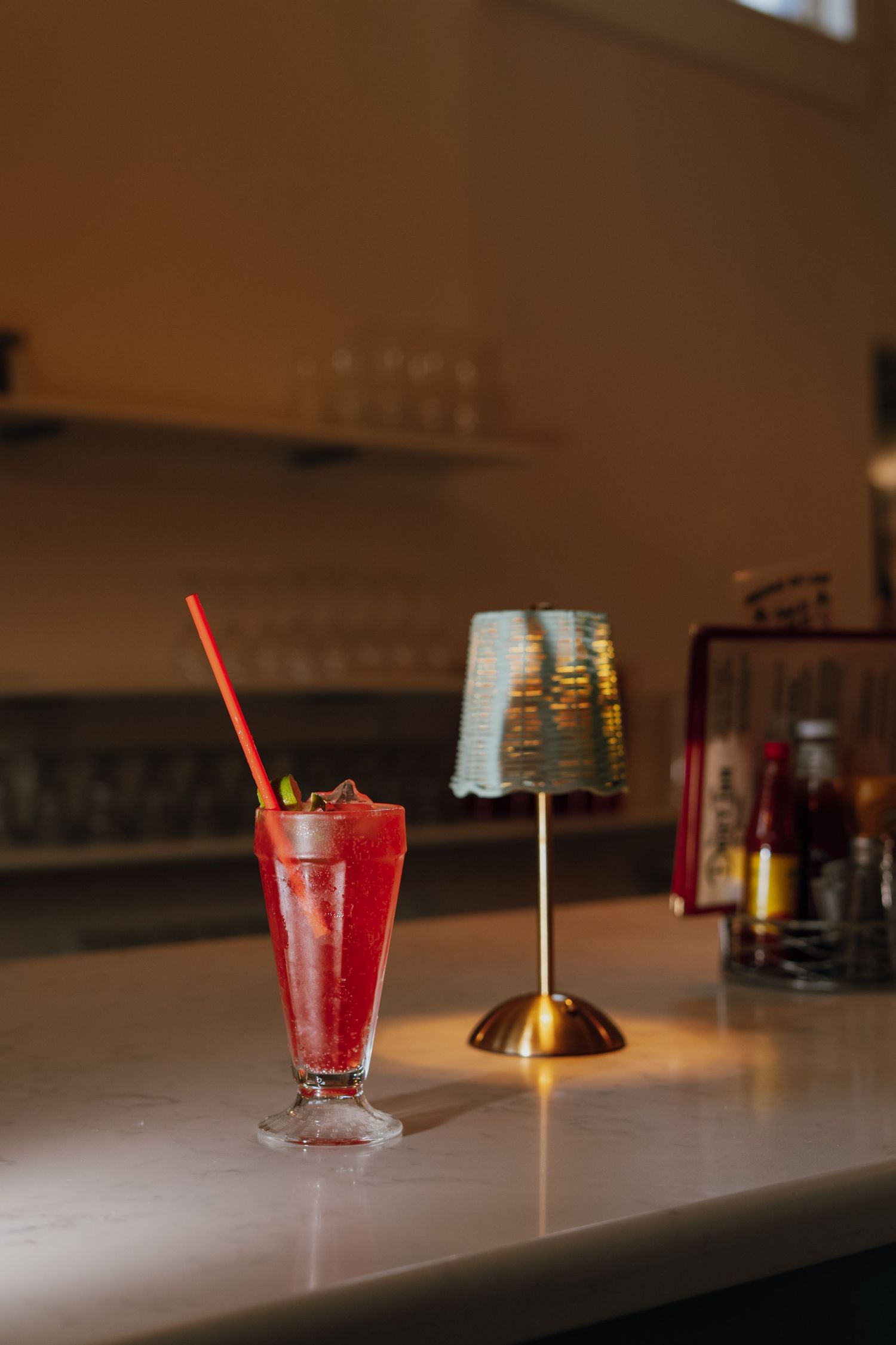 Bright red iced cocktail with straw on white bar counter beside small gold table lamp.