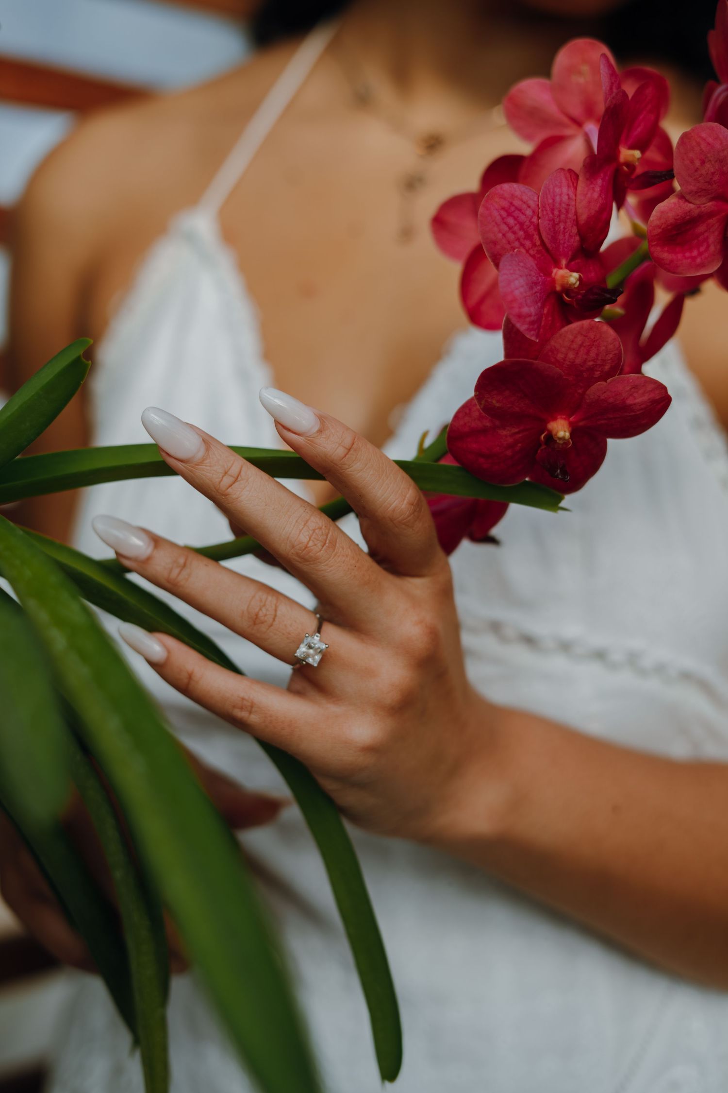 Close-up of a woman's hand with nude nails and diamond ring touching red orchid blooms in white dress.