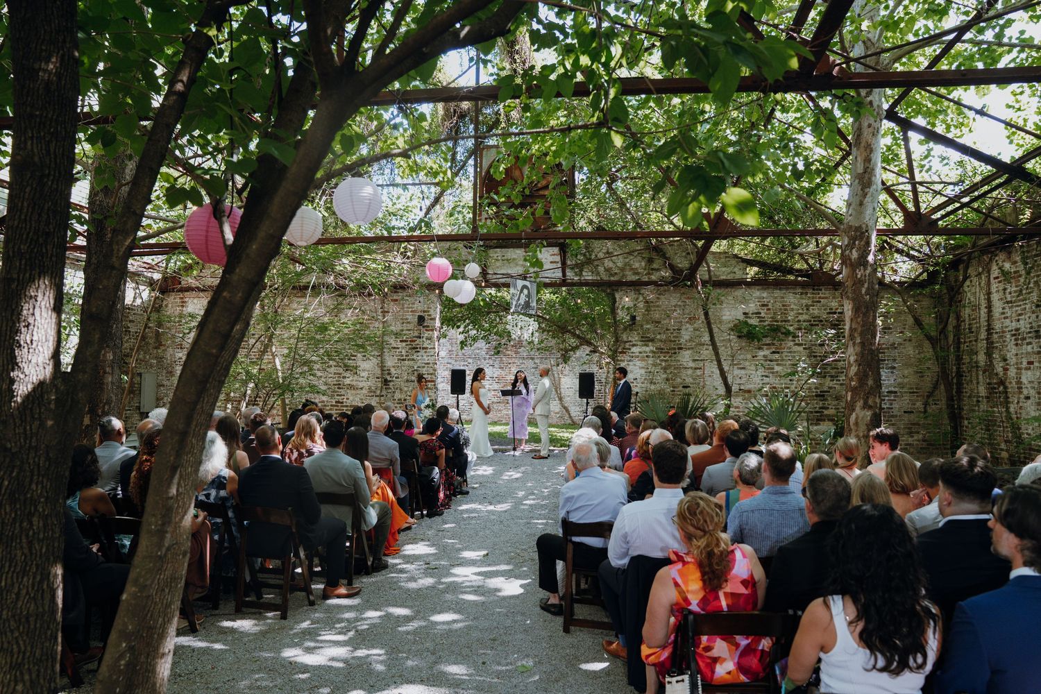 Outdoor wedding ceremony under vine-covered pergola with guests seated along aisle toward altar.