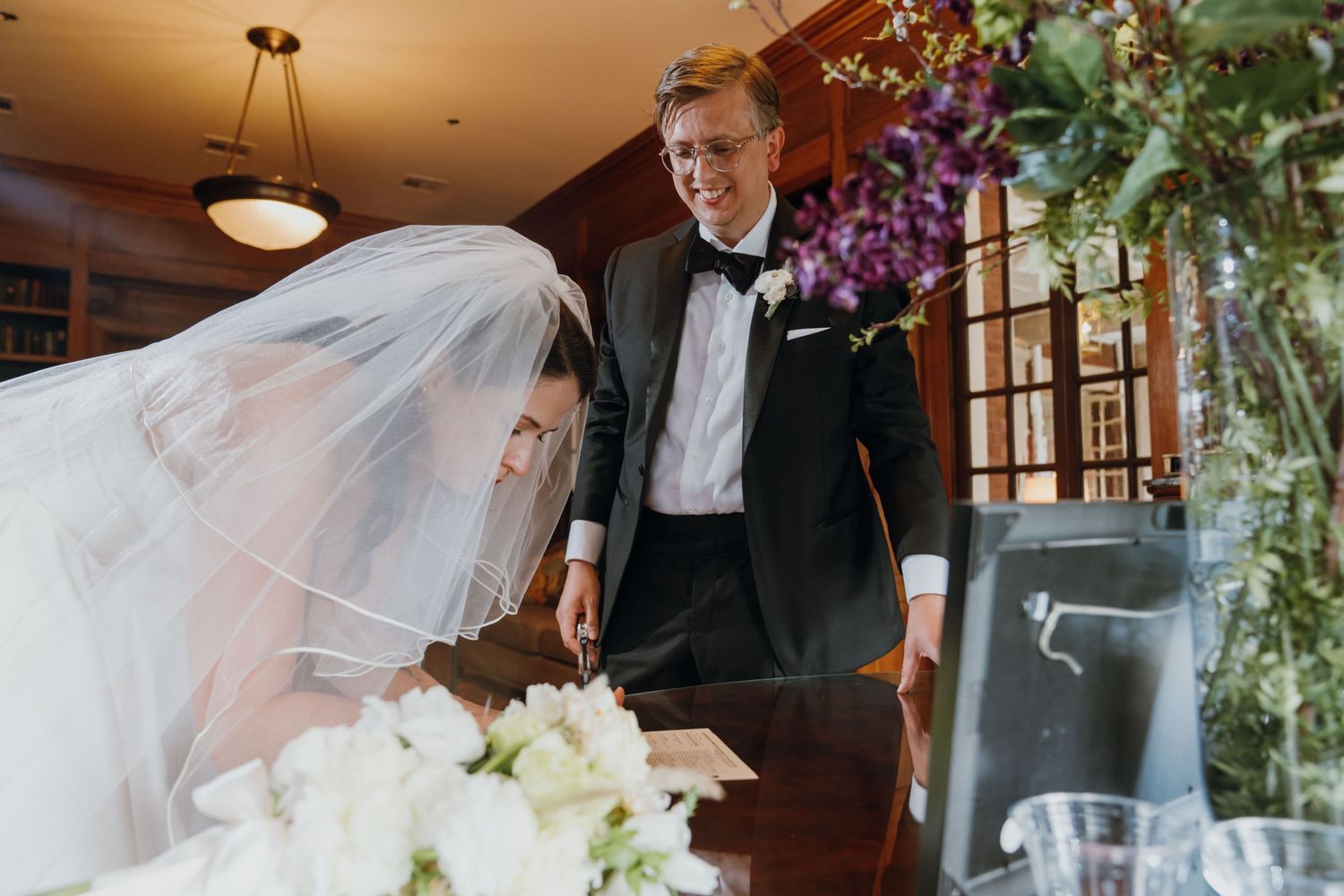 Groom in black tuxedo signing marriage certificate beside veiled bride at decorated table. New Orleans, Louisiana, Mississippi