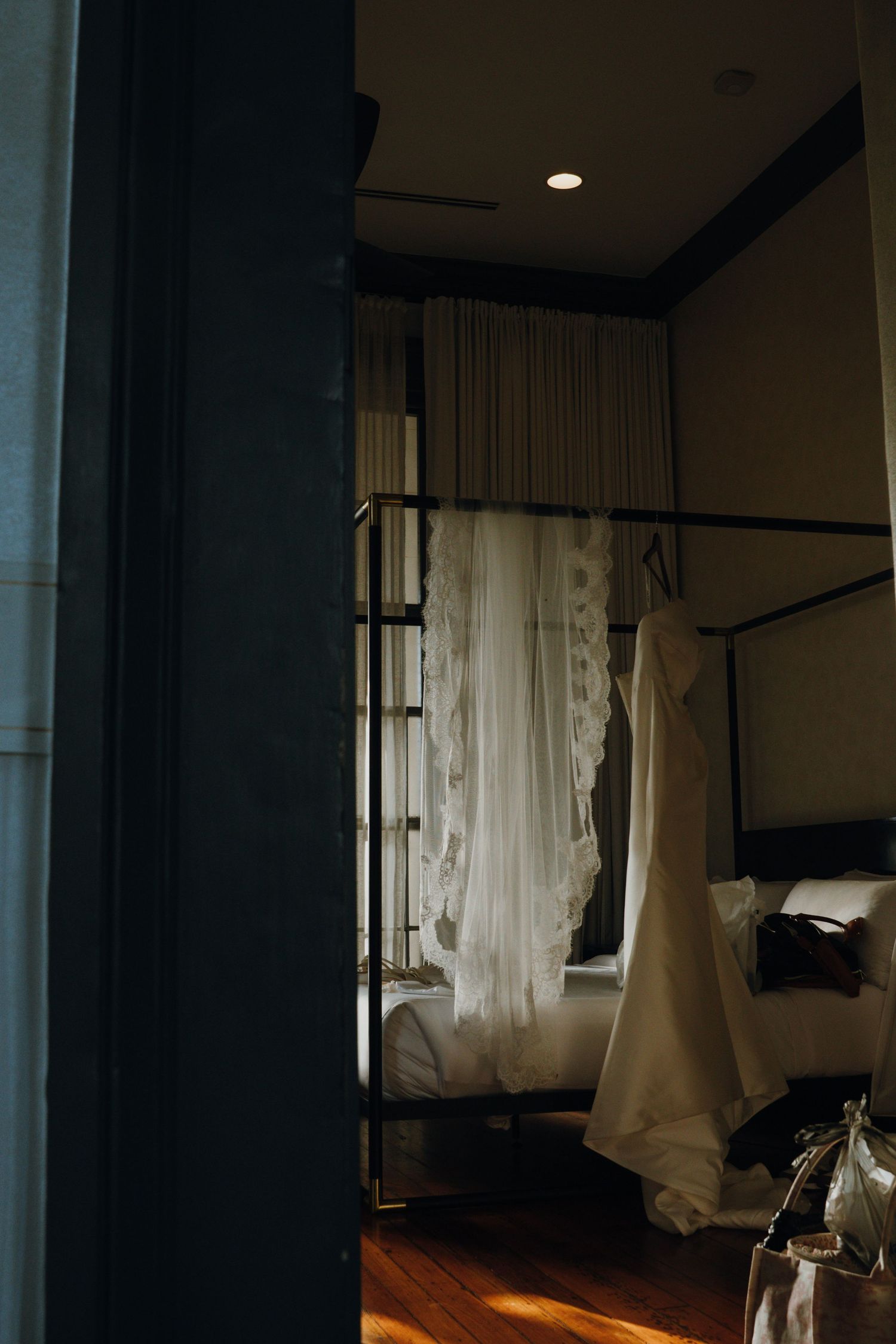 Wedding dress hanging in a dimly lit elegant hotel room with a four-poster bed.