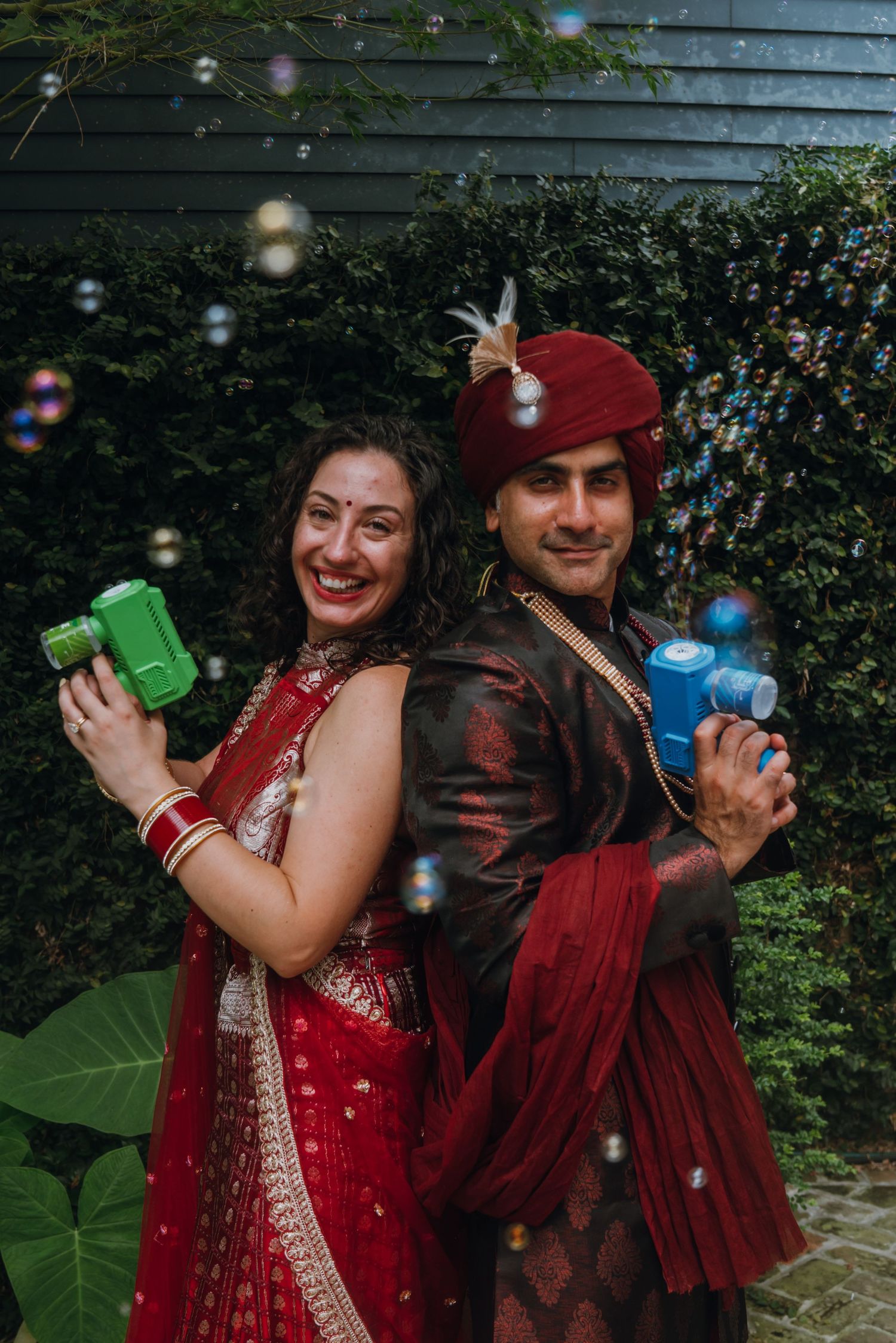 Indian bride and groom posing back-to-back holding colorful bubble guns at their wedding.