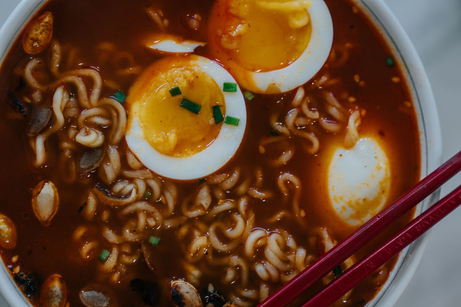 A bowl of spicy ramen soup with soft-boiled eggs, noodles, mushrooms, and green onions, served with red chopsticks.