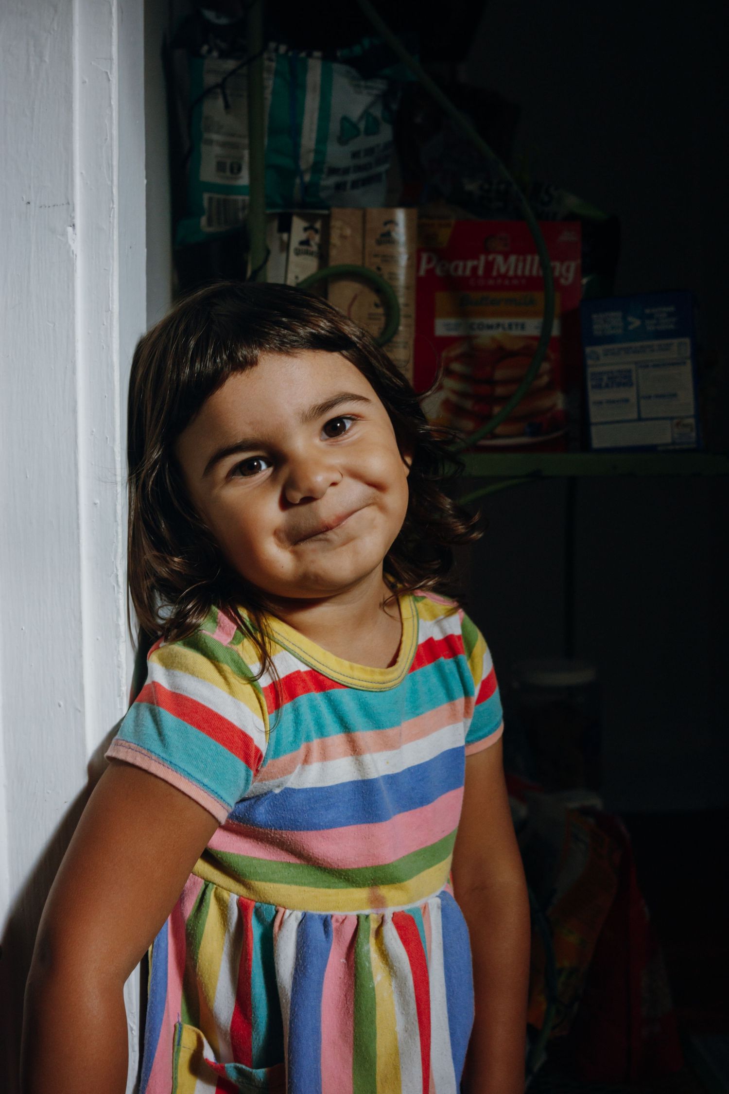 A smiling young girl in a colorful striped dress poses cheerfully indoors with shelves visible behind her.
