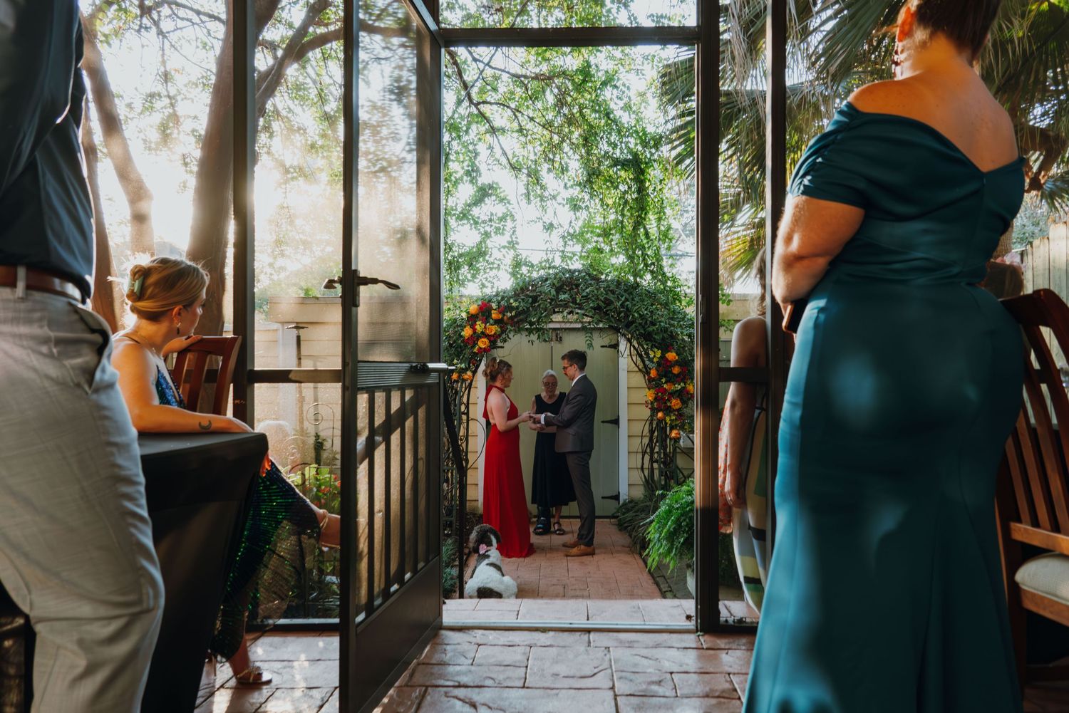Wedding ceremony viewed through glass doors with couple at floral arch, guests watching in elegant venue.