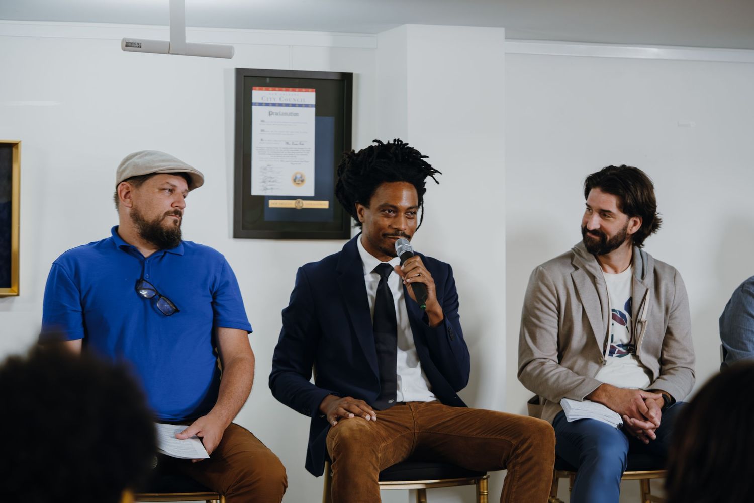 Three men sit in a panel discussion, with the center speaker holding a microphone at an indoor event.