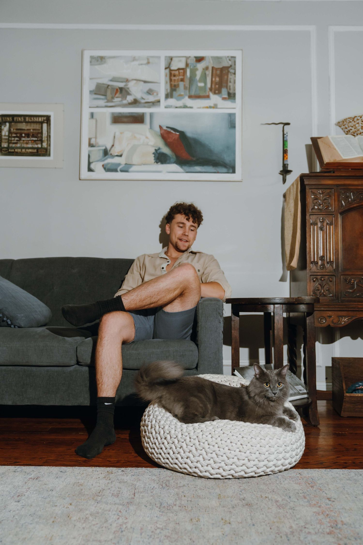Young man sitting on gray sofa smiling, with fluffy gray cat resting on white knitted pouf in cozy living room.