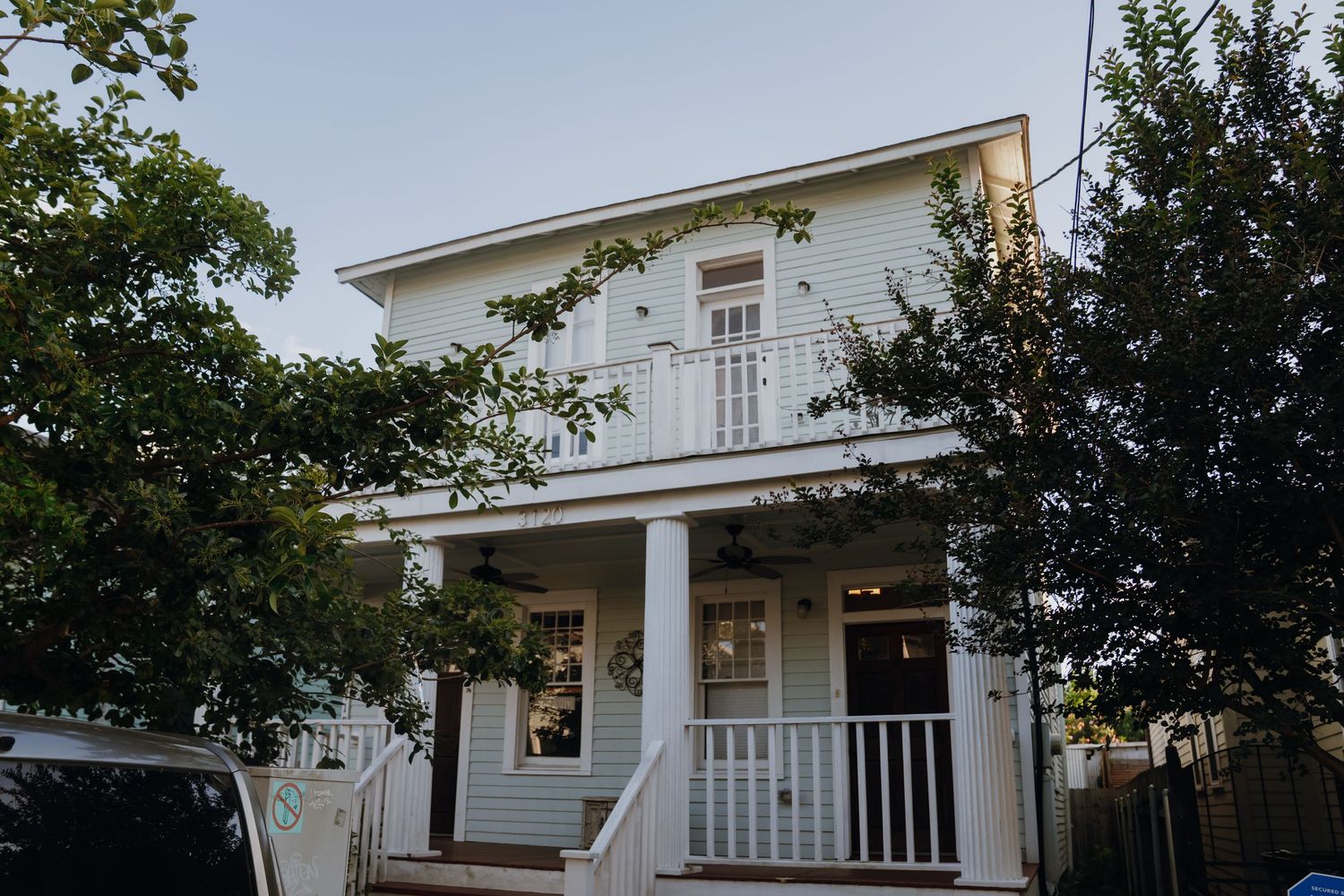 A charming white two-story home with a covered front porch and balcony surrounded by lush trees.