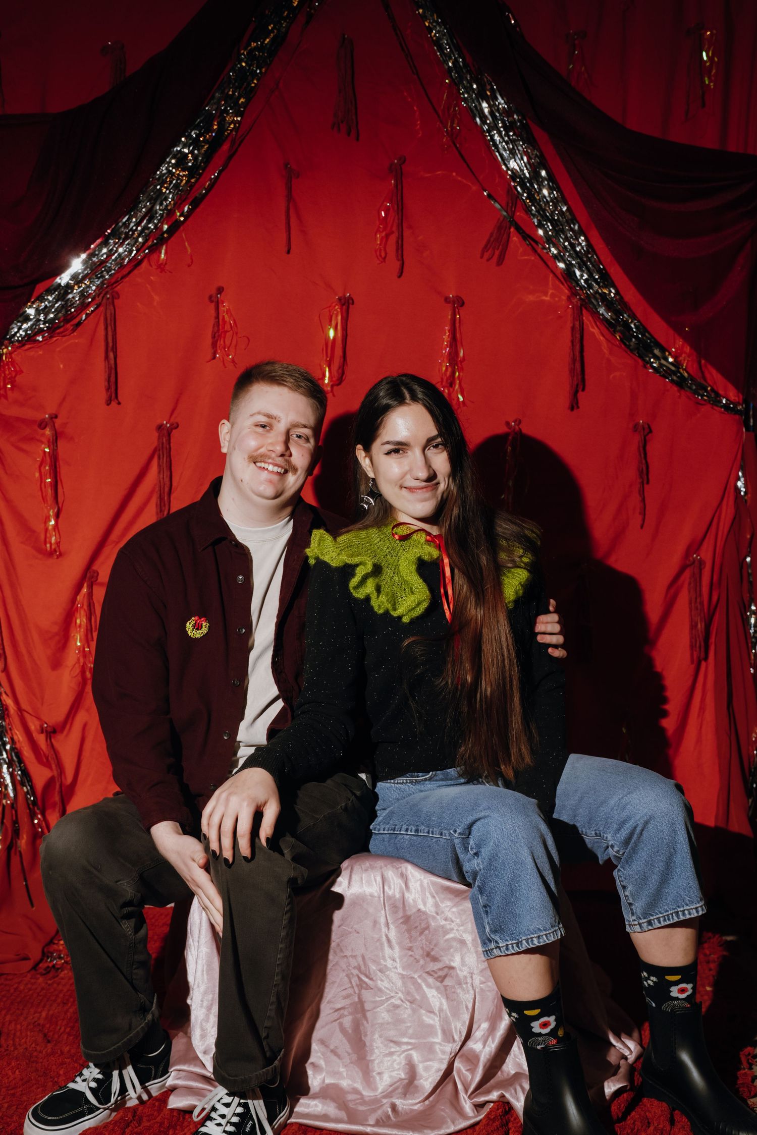 Two smiling people pose together on a pink satin bench against a festive red curtain backdrop with gold tinsel.