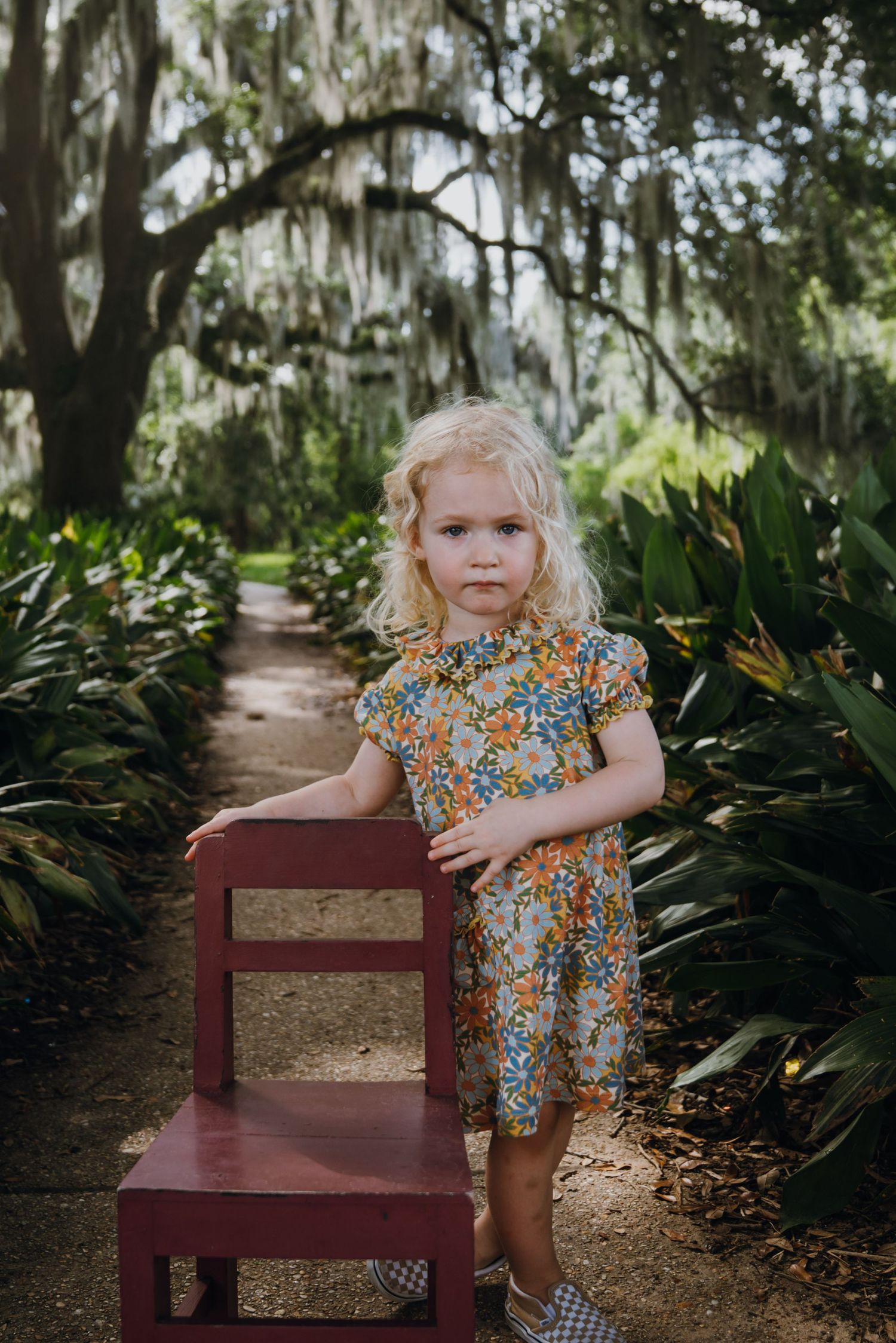 Blonde toddler girl in floral dress standing beside red chair on garden path with Spanish moss trees.