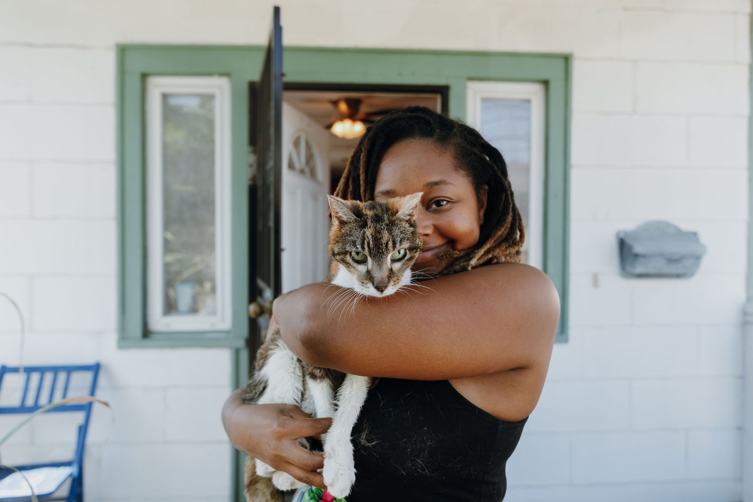 A smiling woman hugging a tabby cat in front of a white house with a green door.