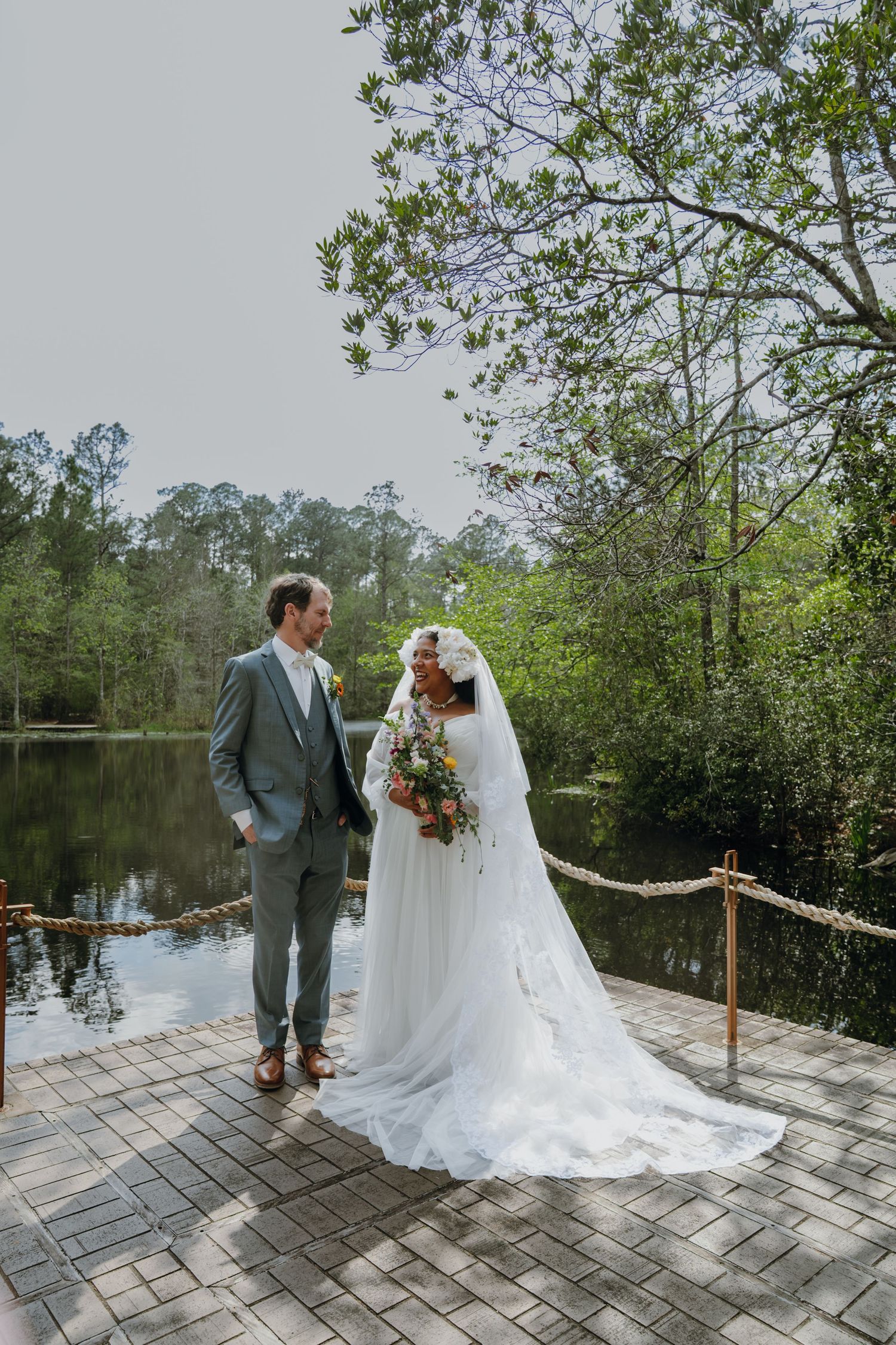 Bride and groom pose on wooden dock surrounded by serene lake and forest.