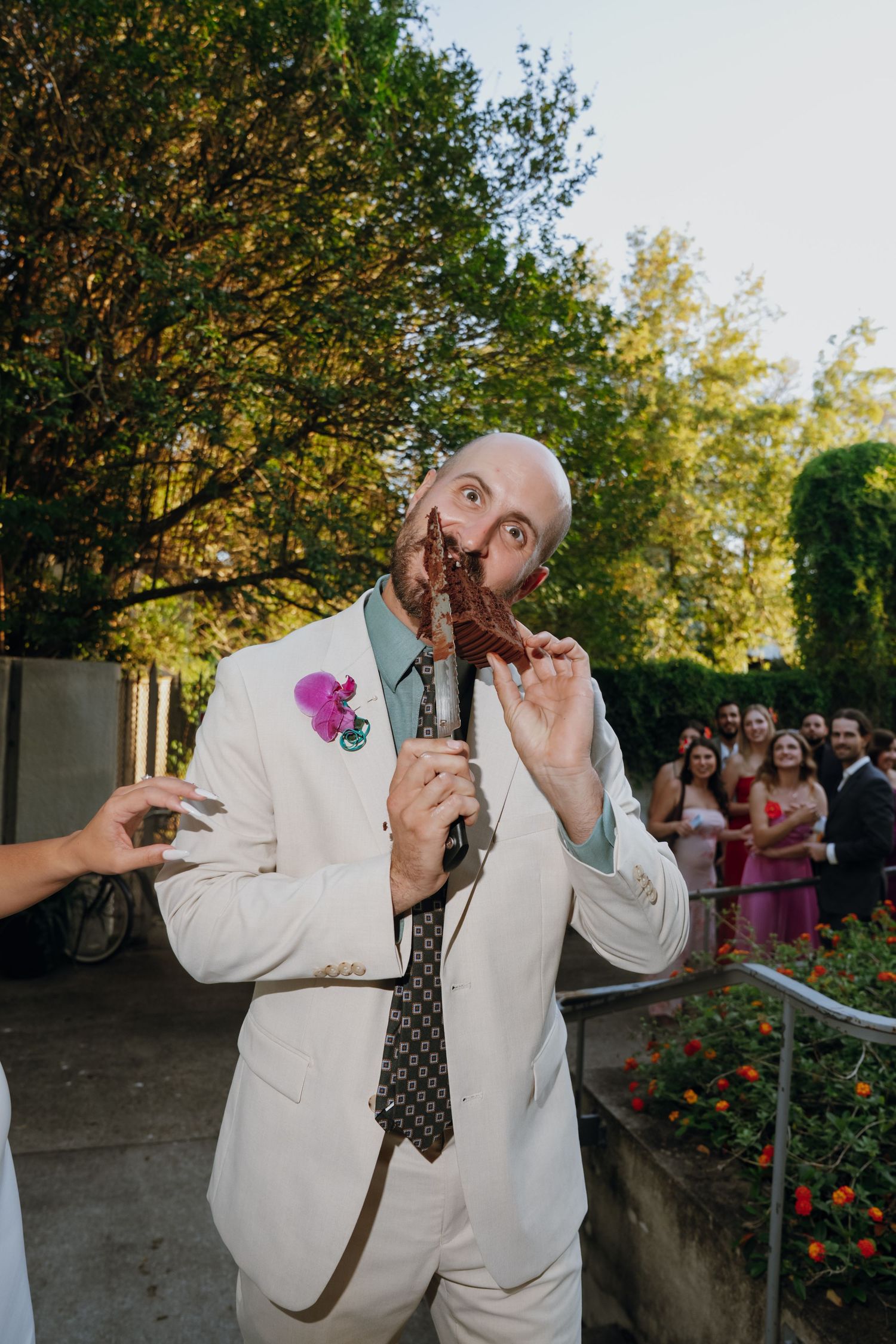 Bald groom in white suit with pink boutonniere playfully poses with decorative item at outdoor wedding reception.