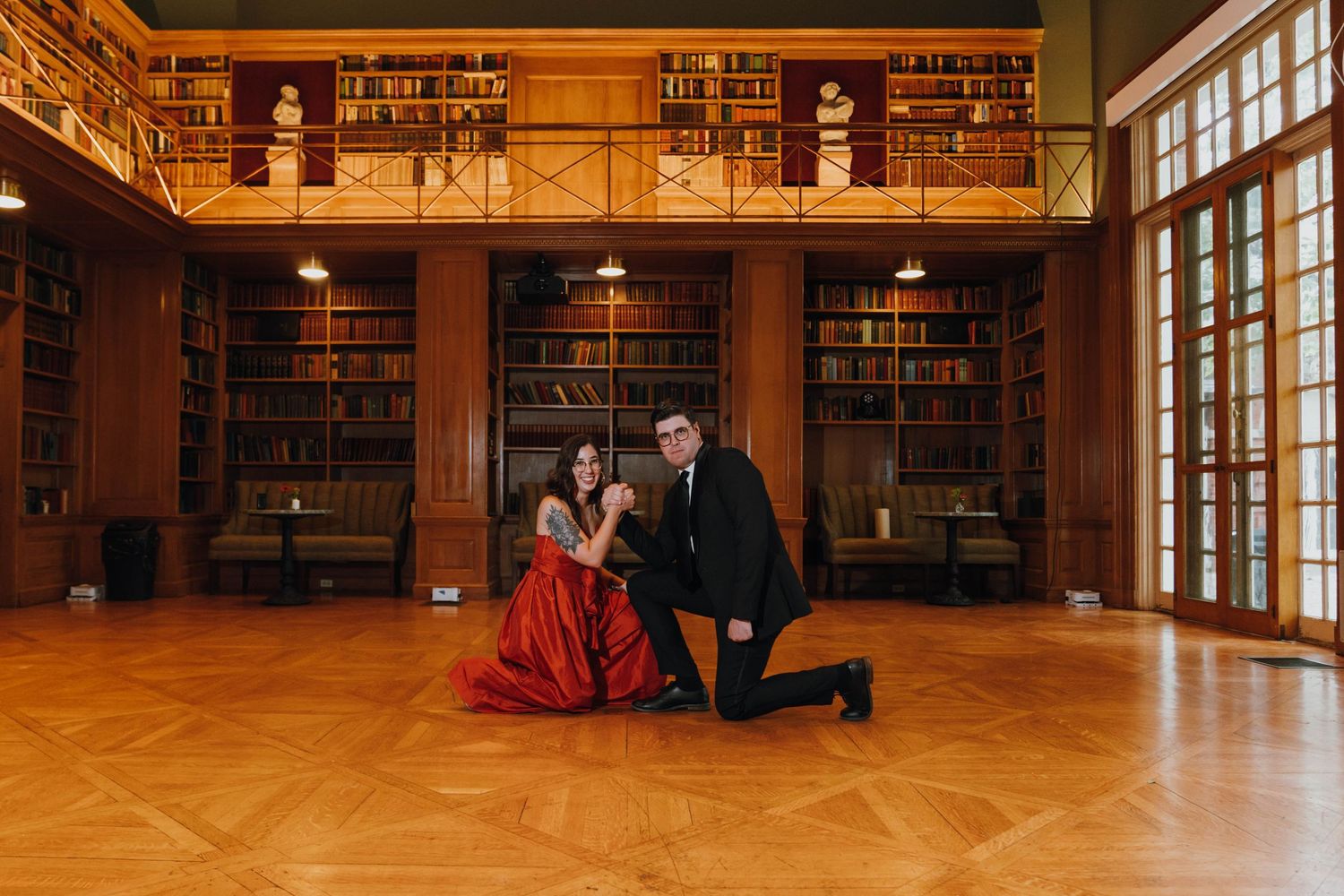 Couple poses dramatically in a grand library with floor-to-ceiling bookshelves, bride wearing a red gown. New Orleans, Louisiana, Mississipp