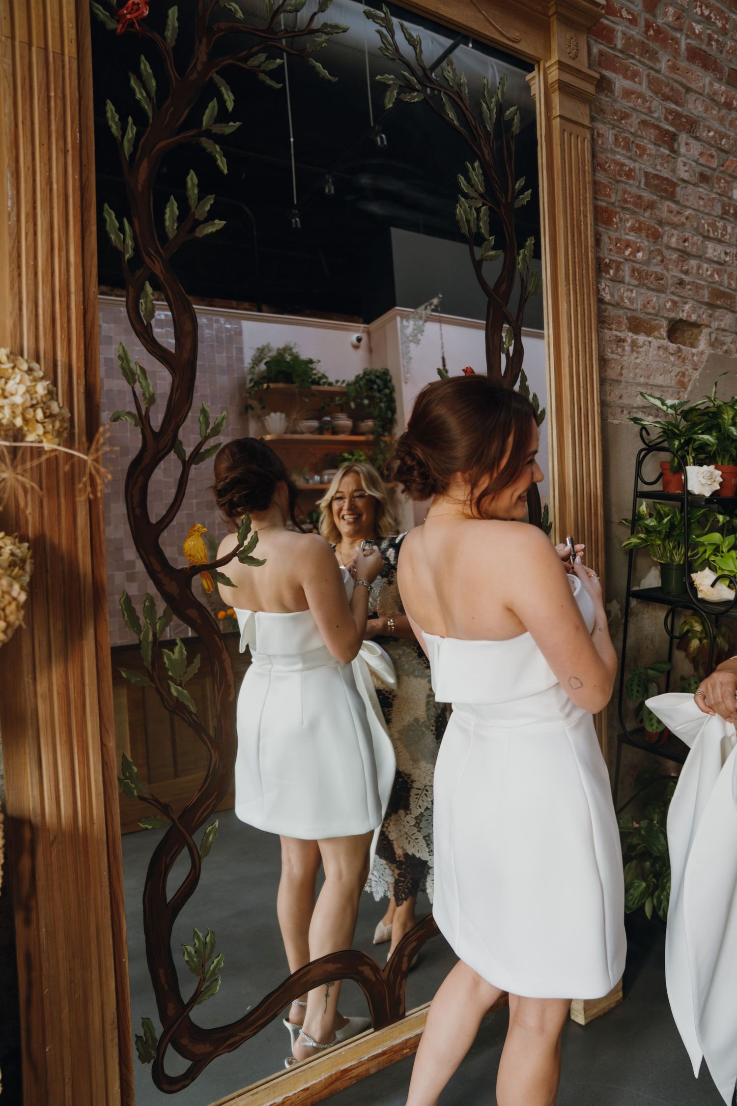 Bride in white mini dress viewing reflection in ornate tree-framed mirror at floral shop.