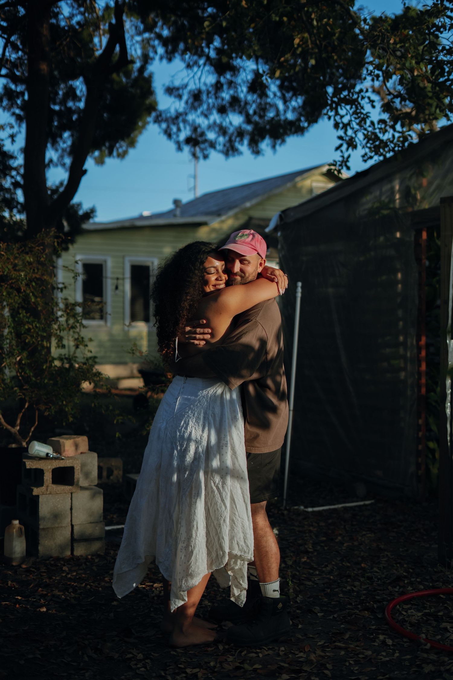 A couple hugs tenderly in a sunlit backyard with a greenhouse and house visible in the background.