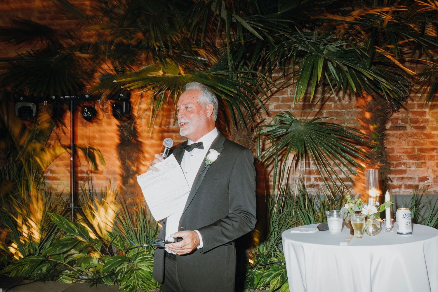Man in tuxedo delivering wedding speech at elegant reception with tropical greenery backdrop.